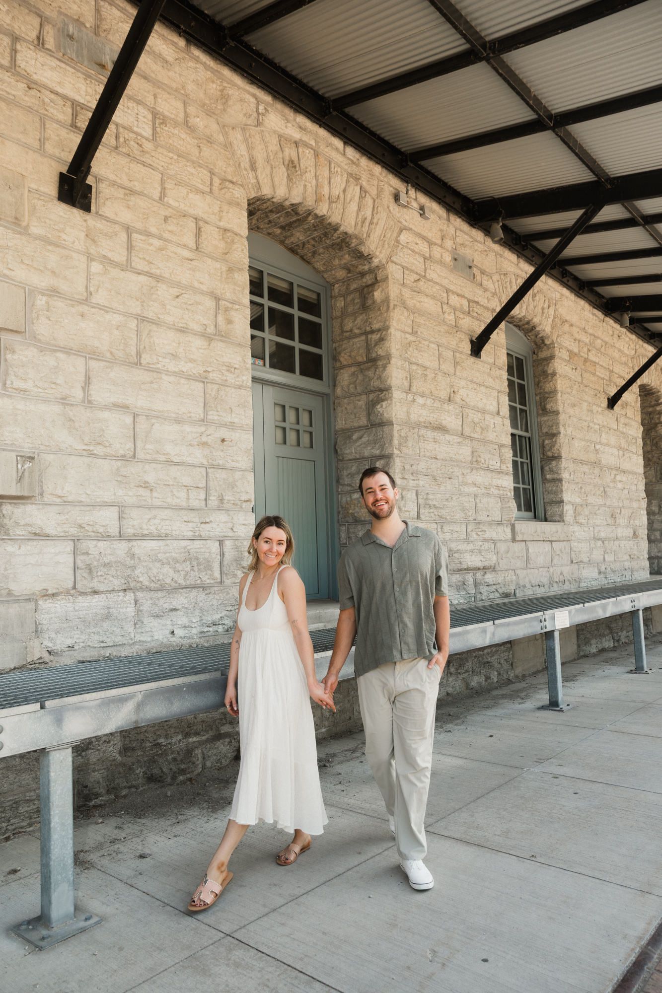 Couple holding hands and smiling in front of a historic stone building, woman in white dress, man in gray shirt.