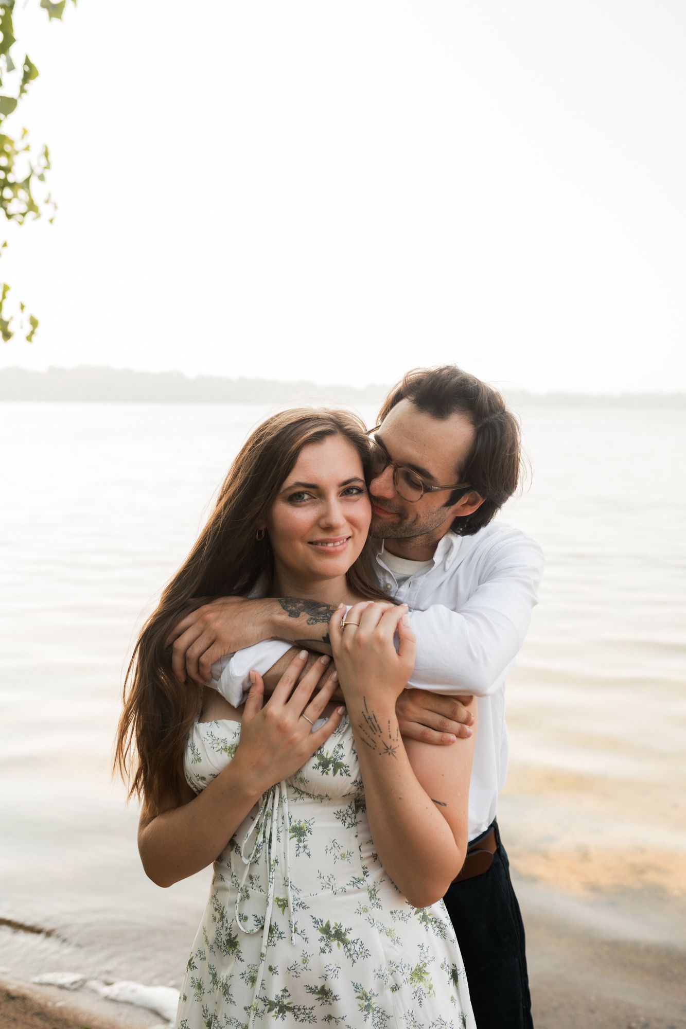 Couple embracing by a serene lake at sunset, woman in floral dress smiling as man hugs her from behind.