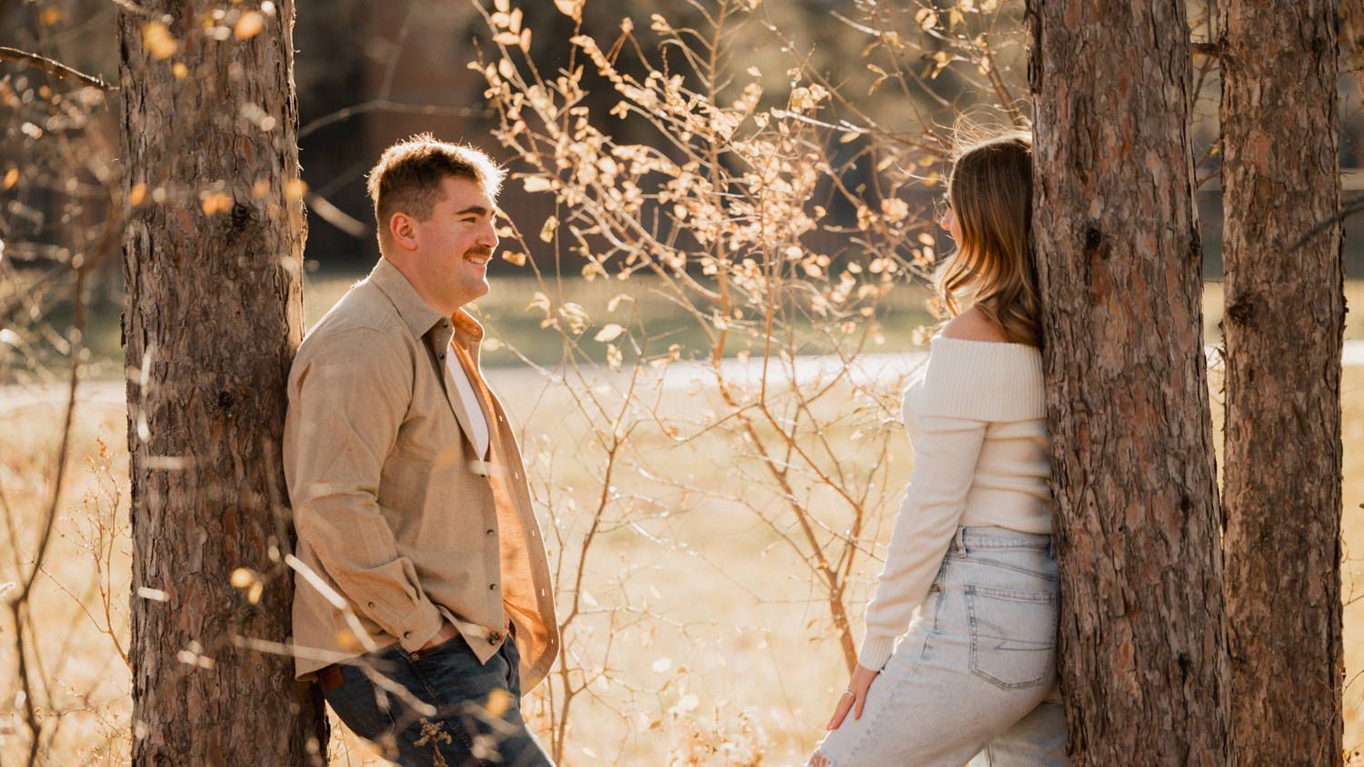 A couple shares affectionate moments between tall pine trees in golden afternoon light.