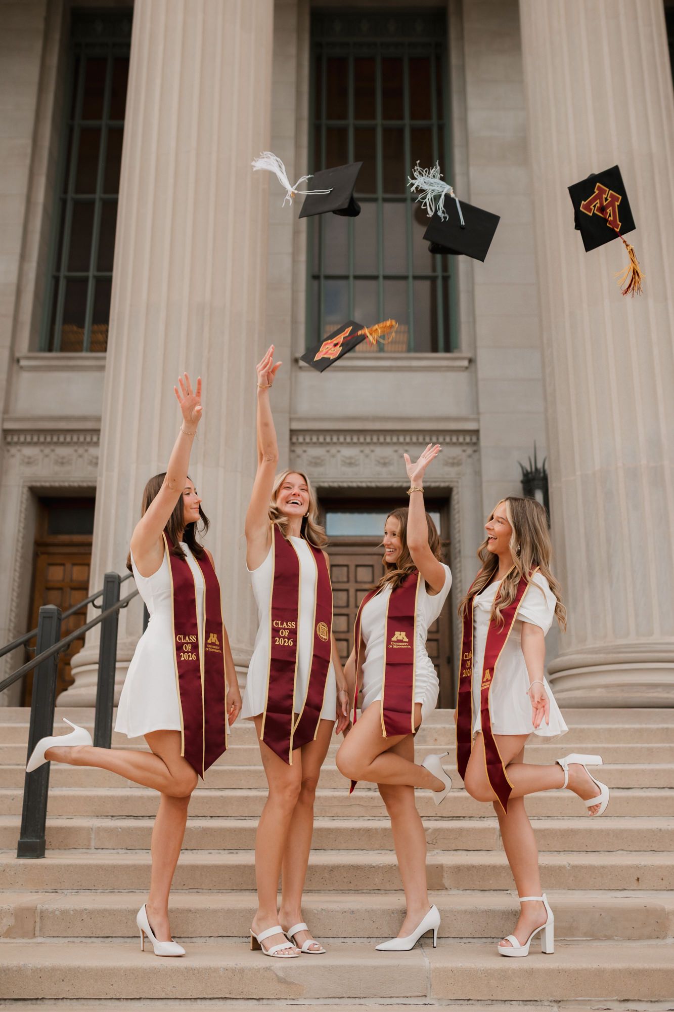 Four University of Minnesota graduates in white dresses toss caps in the air on building steps.