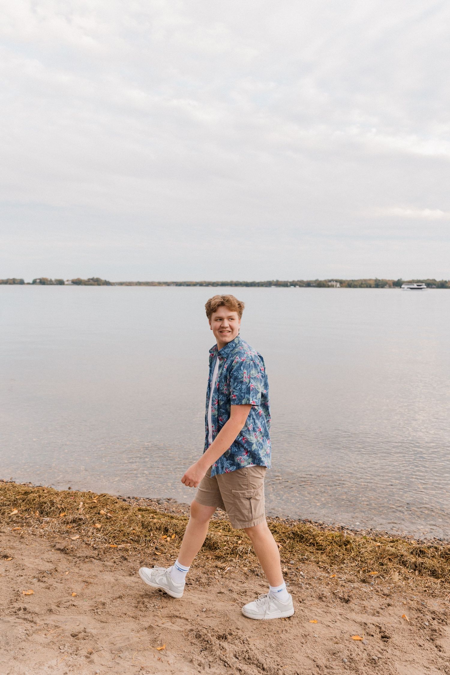 Person in a blue patterned shirt and khaki shorts walking along a calm lakeshore on an overcast day.