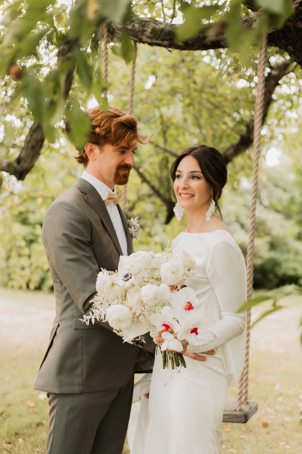 A romantic outdoor wedding photoshoot with a couple posing near a tree with white floral bouquet.