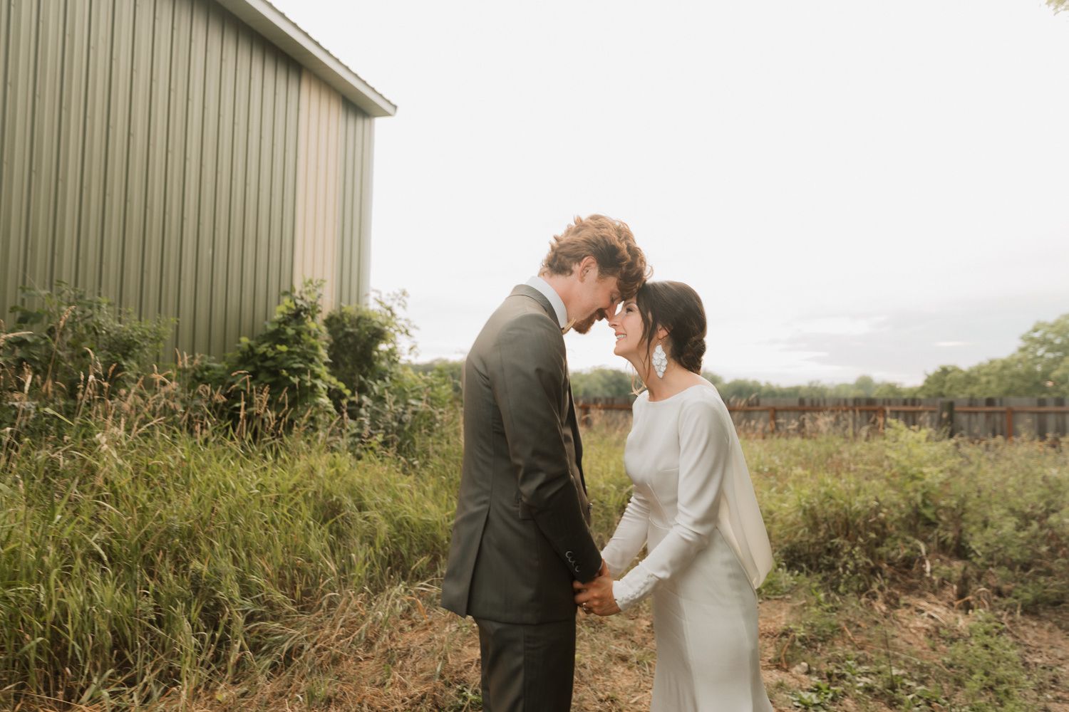 Couple shares intimate moment in front of rustic barn surrounded by tall grass and natural scenery.