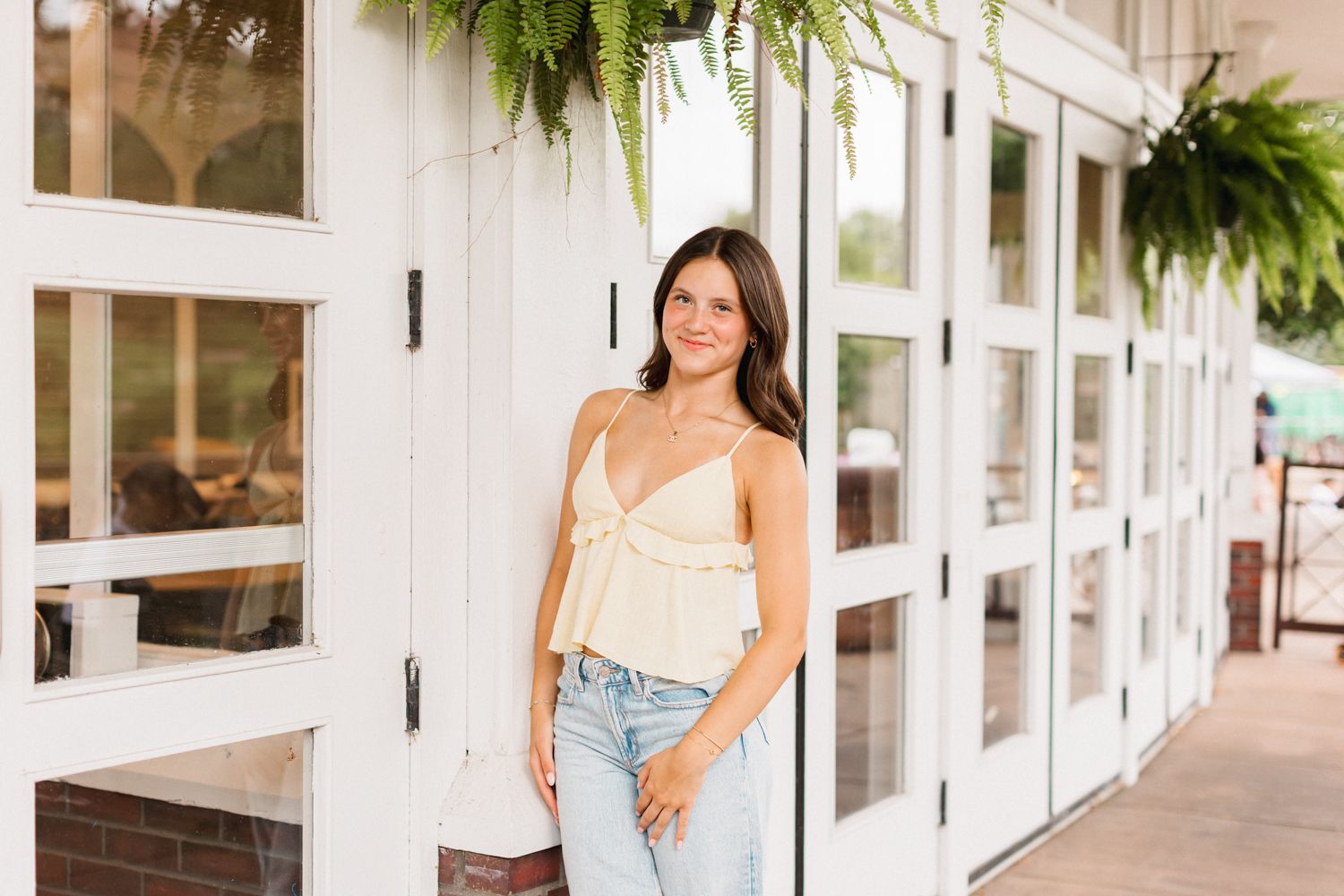 Woman in white dress walks along covered outdoor walkway with hanging ferns and white columns.