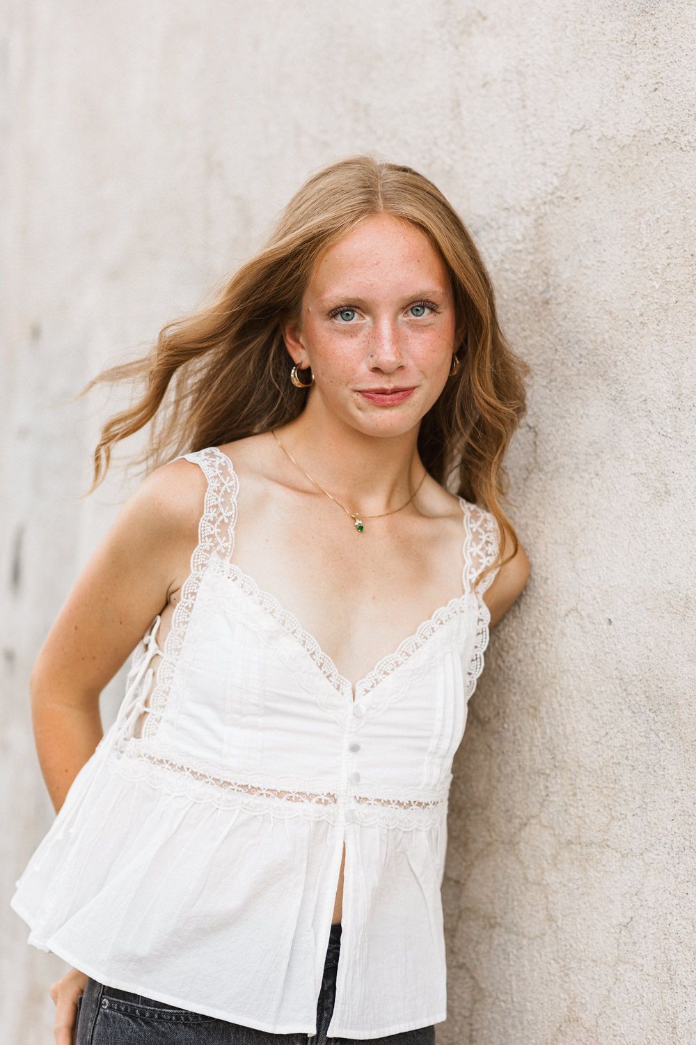 A person in a white lace-trimmed camisole top poses against a light colored wall with hair gently blowing in the breeze.