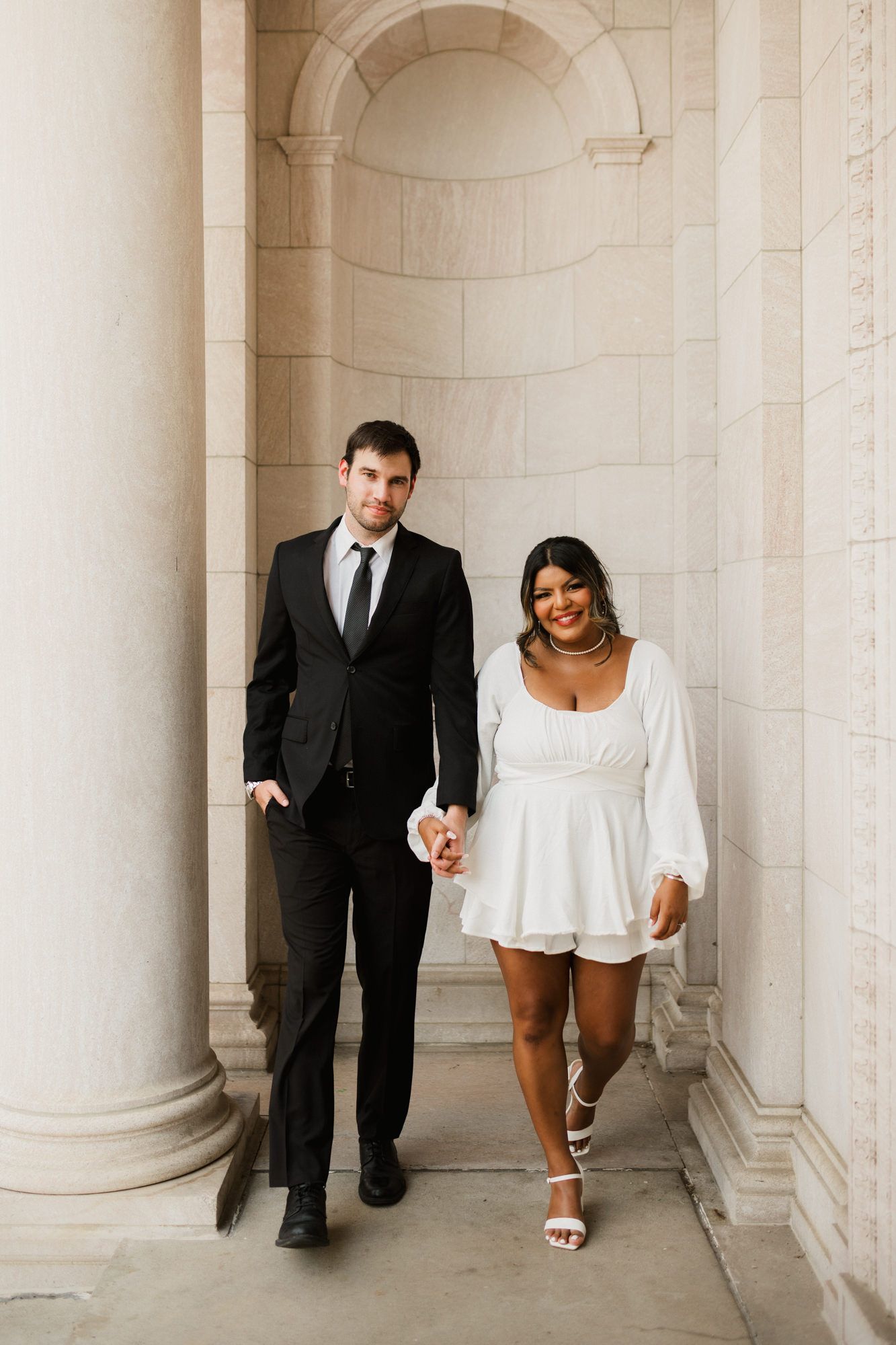 Couple holding hands walking through stone columns, man in black suit, woman in white mini dress and heels.