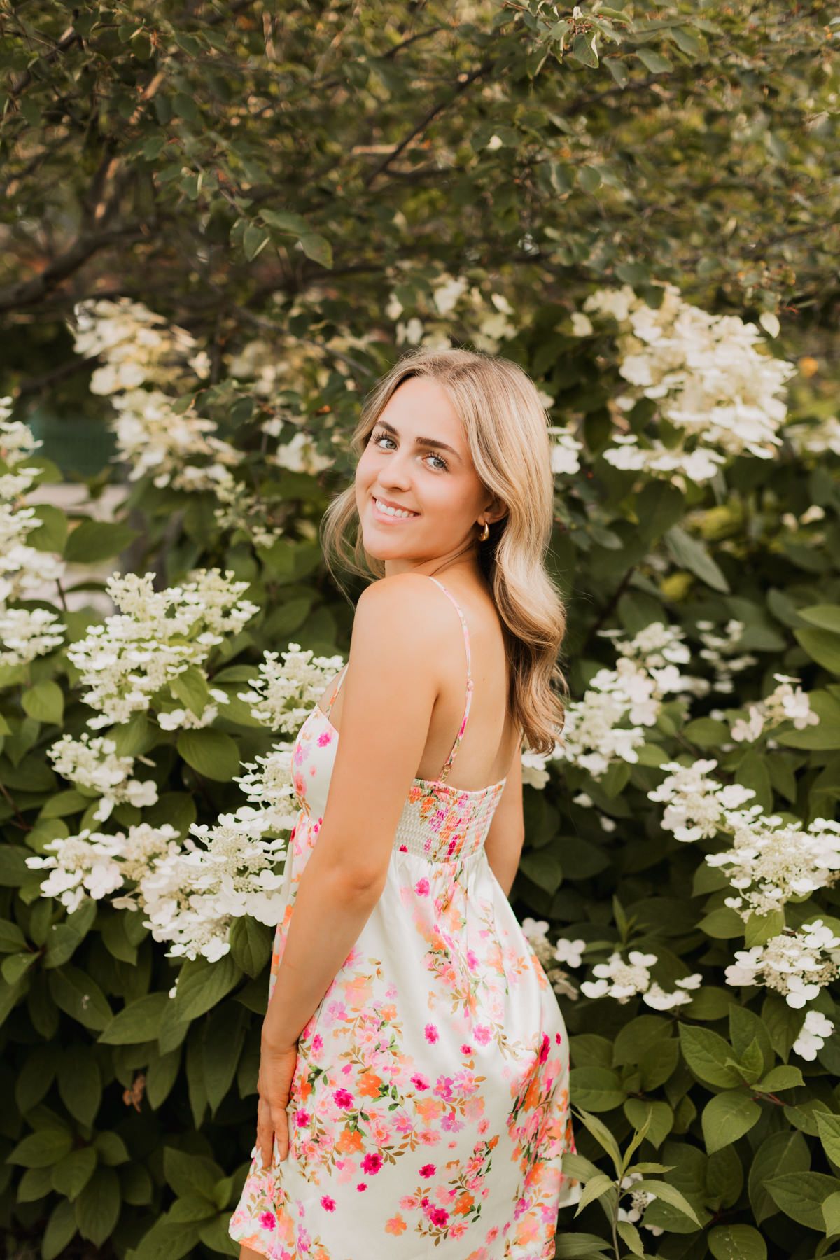 Blonde woman in colorful floral sundress smiles over her shoulder surrounded by blooming white hydrangea bushes.
