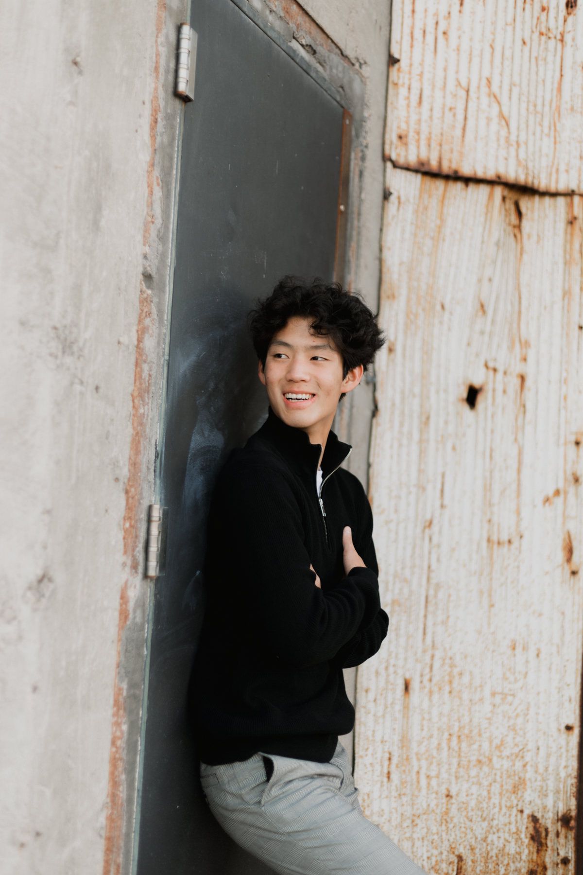 Smiling young Asian man in black zip-up jacket leaning against rustic metal door with arms crossed.