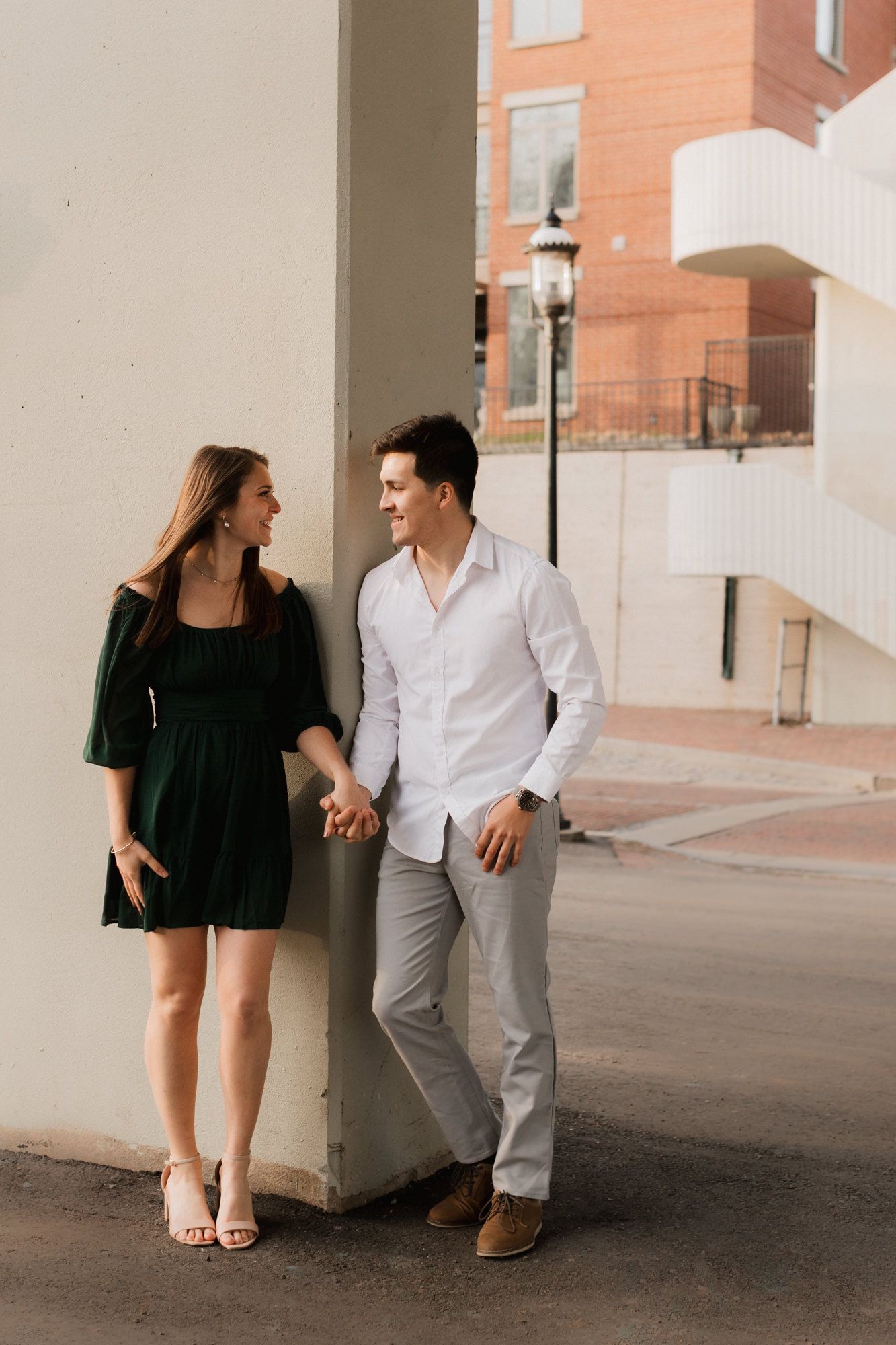 A couple holds hands and smiles at each other while leaning against a pillar in an urban setting.