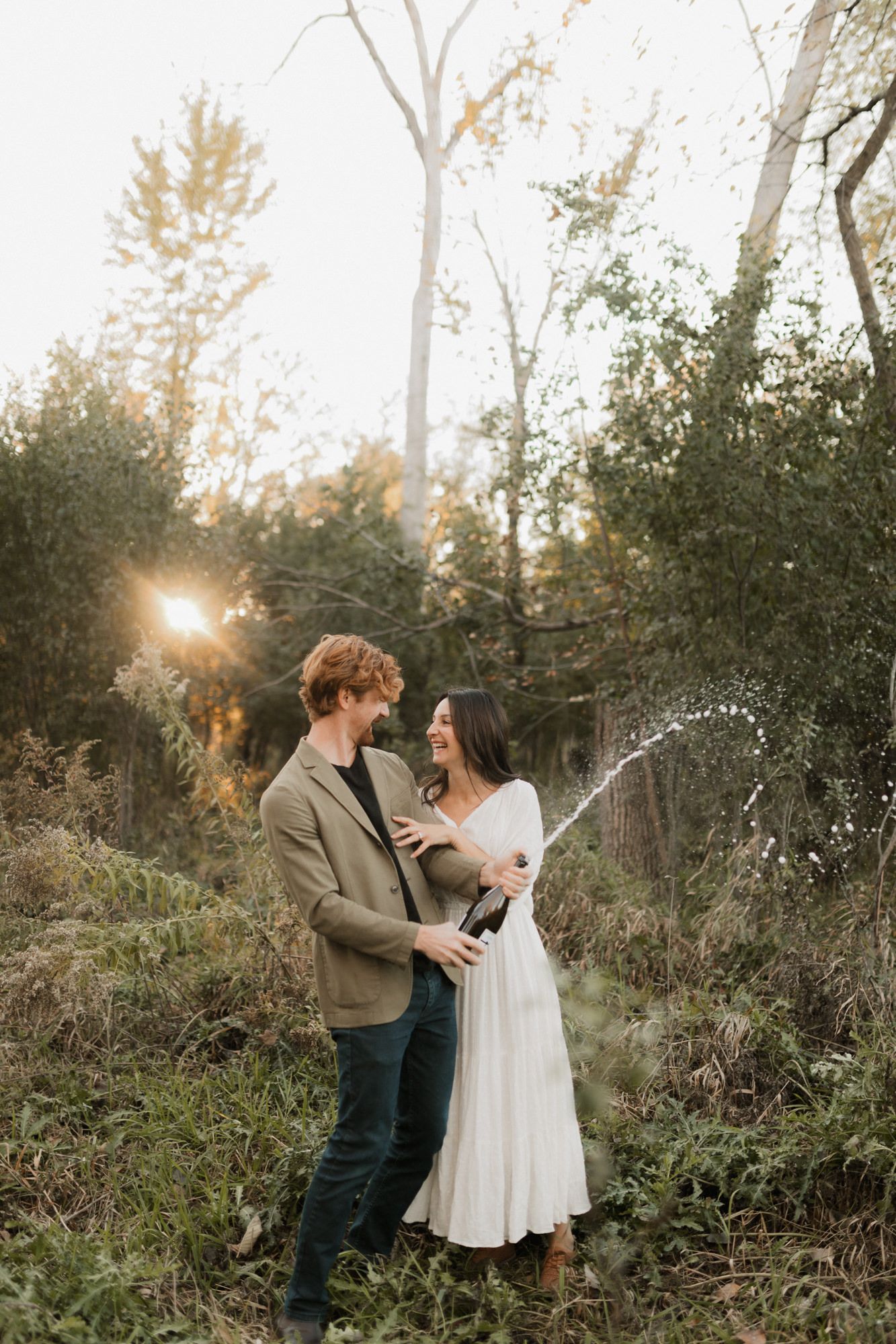 Joyful engaged couple popping champagne at golden hour in a lush forest setting.