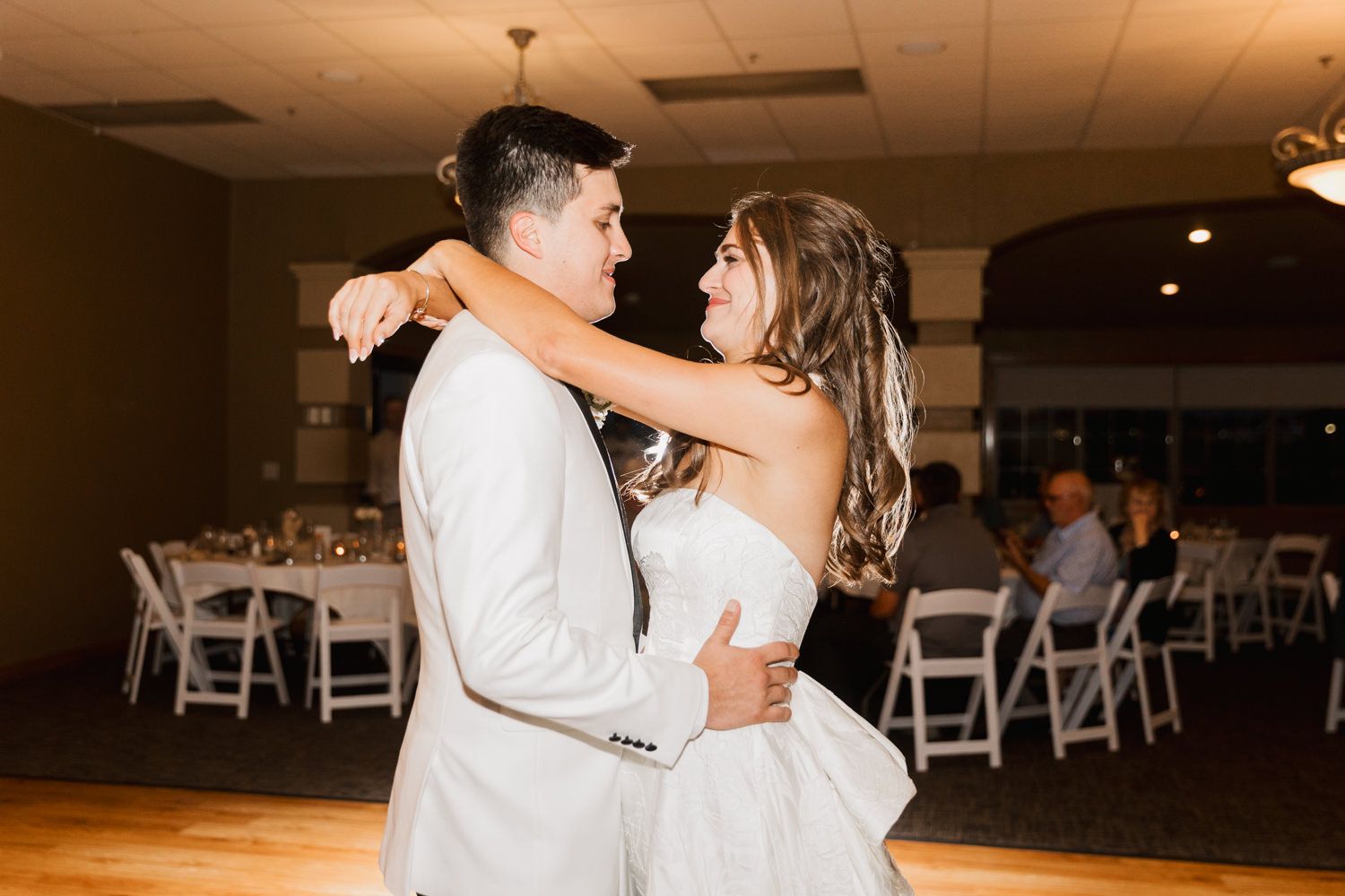 A couple dances together on a wooden dance floor in a reception hall with white chairs visible in the background.