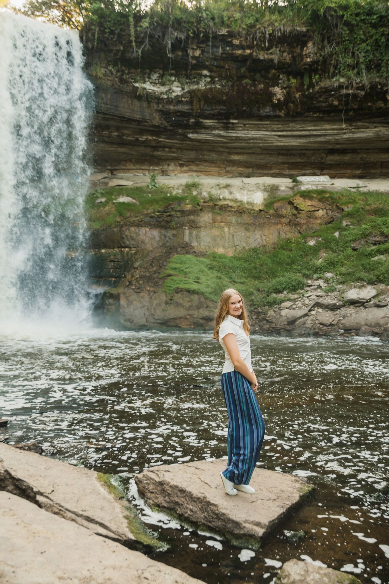 Young woman in blue pants and white top smiling near a waterfall, standing on rocks with greenery in background.