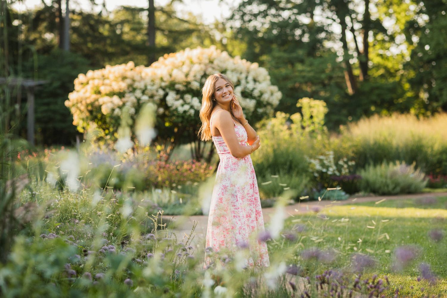 A young woman in a floral pink dress poses gracefully among lush garden blooms in warm golden sunlight.