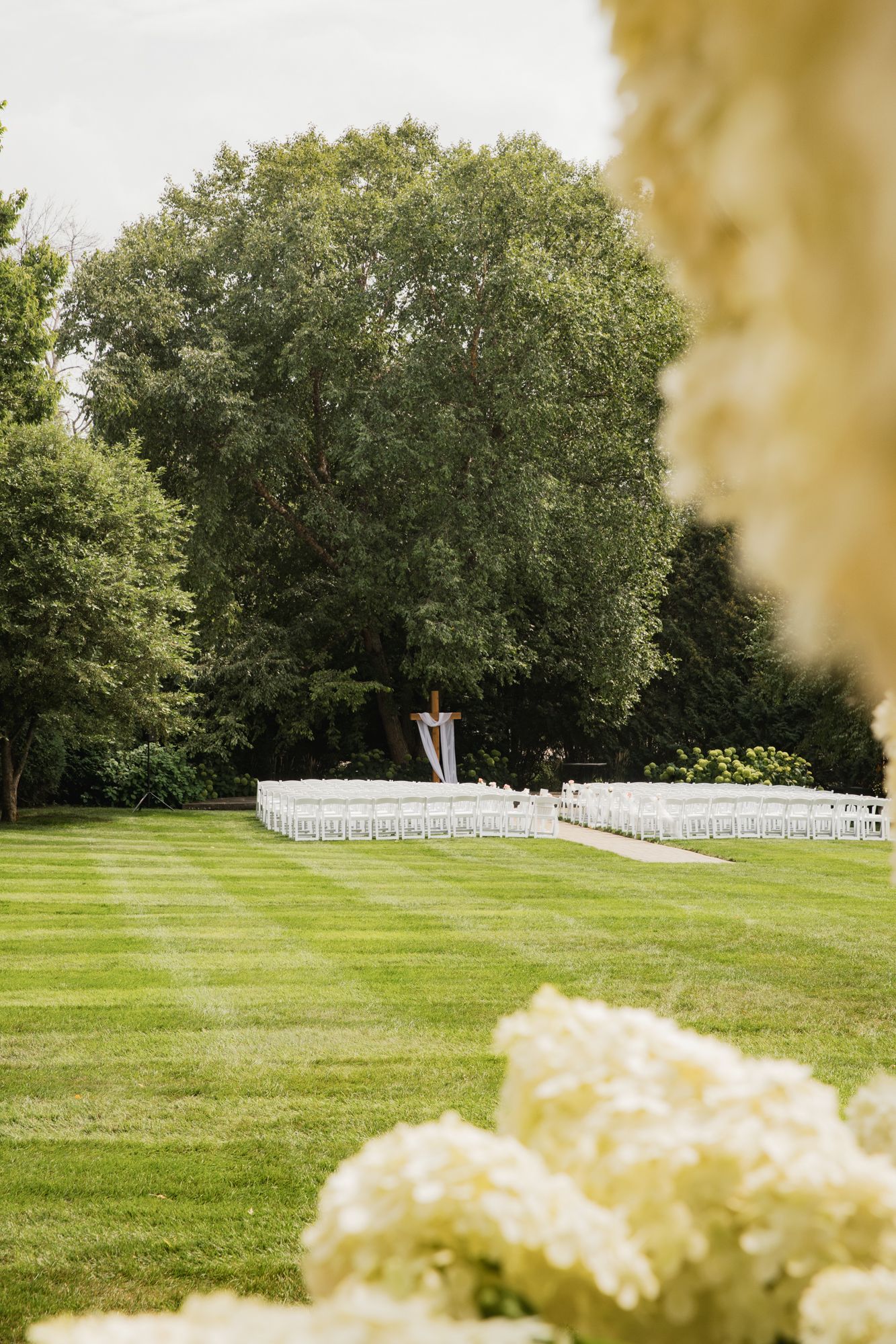 Outdoor wedding ceremony setup with white chairs on striped green lawn surrounded by lush trees and white hydrangeas in foreground.