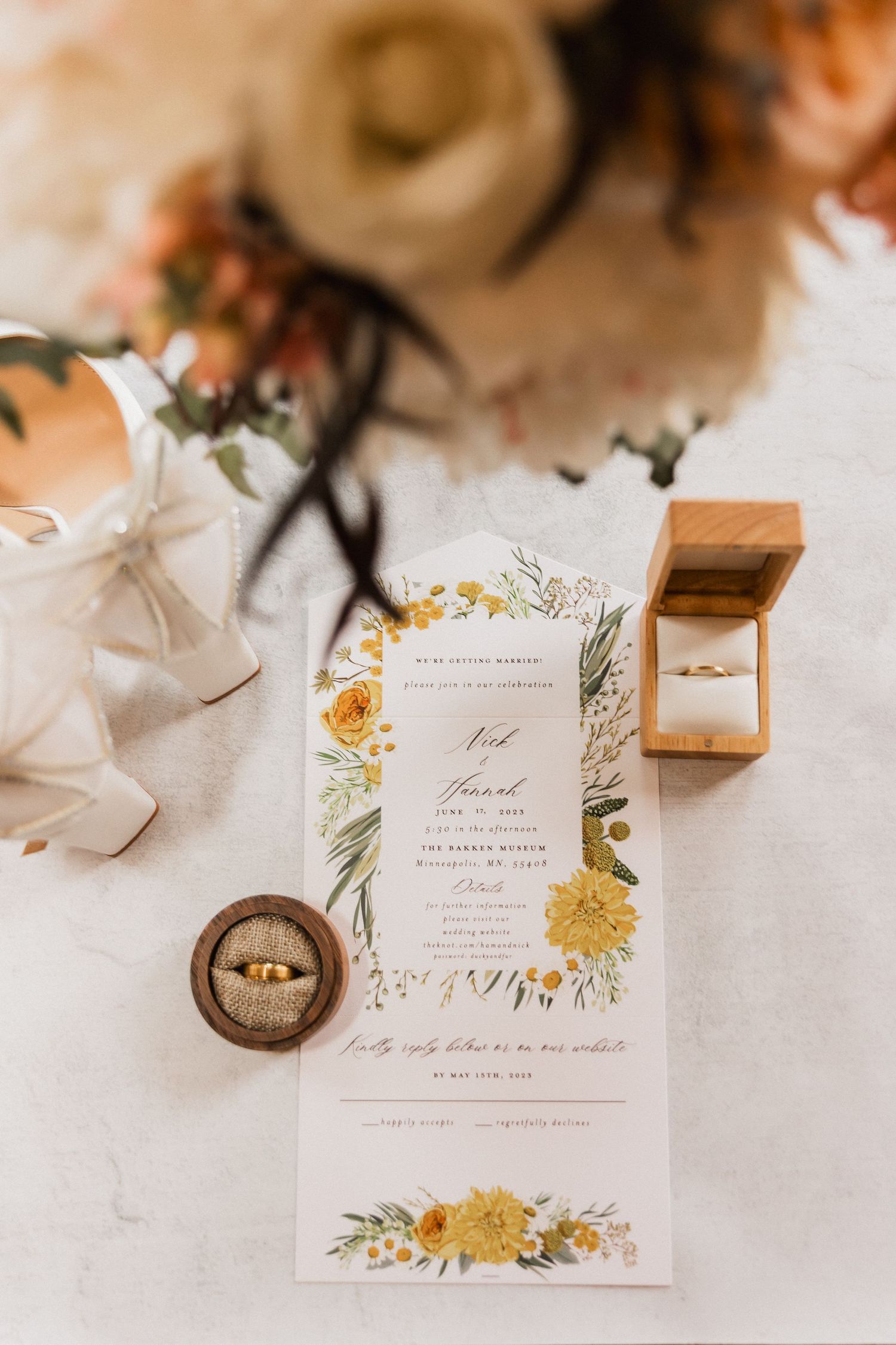 Elegant wedding invitation with yellow floral details, gold rings in wooden box, white heels, and bouquet on marble surface.