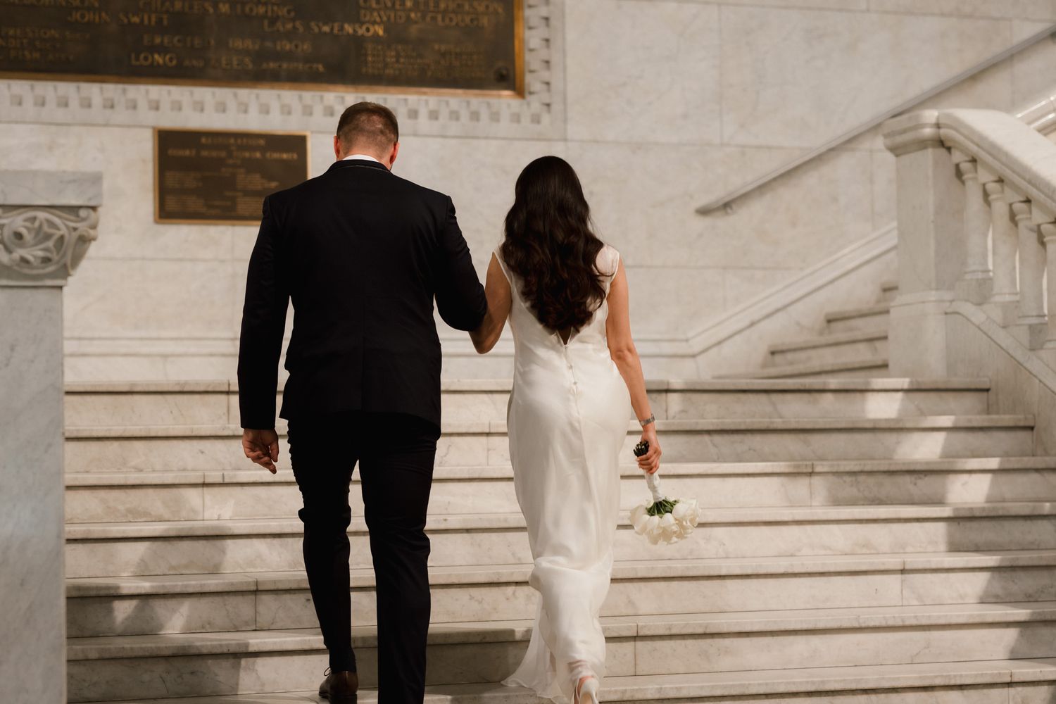 Bride and groom walking up marble stairs holding hands, bride in white gown holding bouquet, groom in black suit.