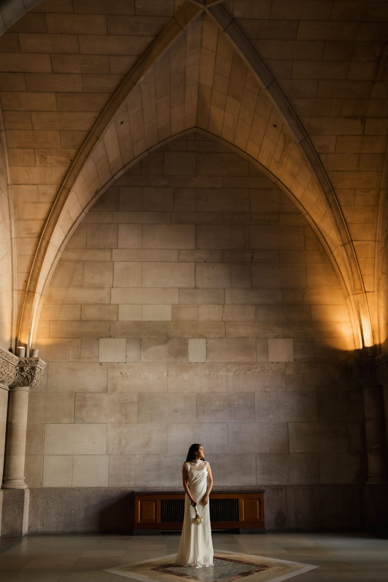 Bride in white gown stands beneath Gothic stone arch in grand cathedral hall with warm ambient lighting.