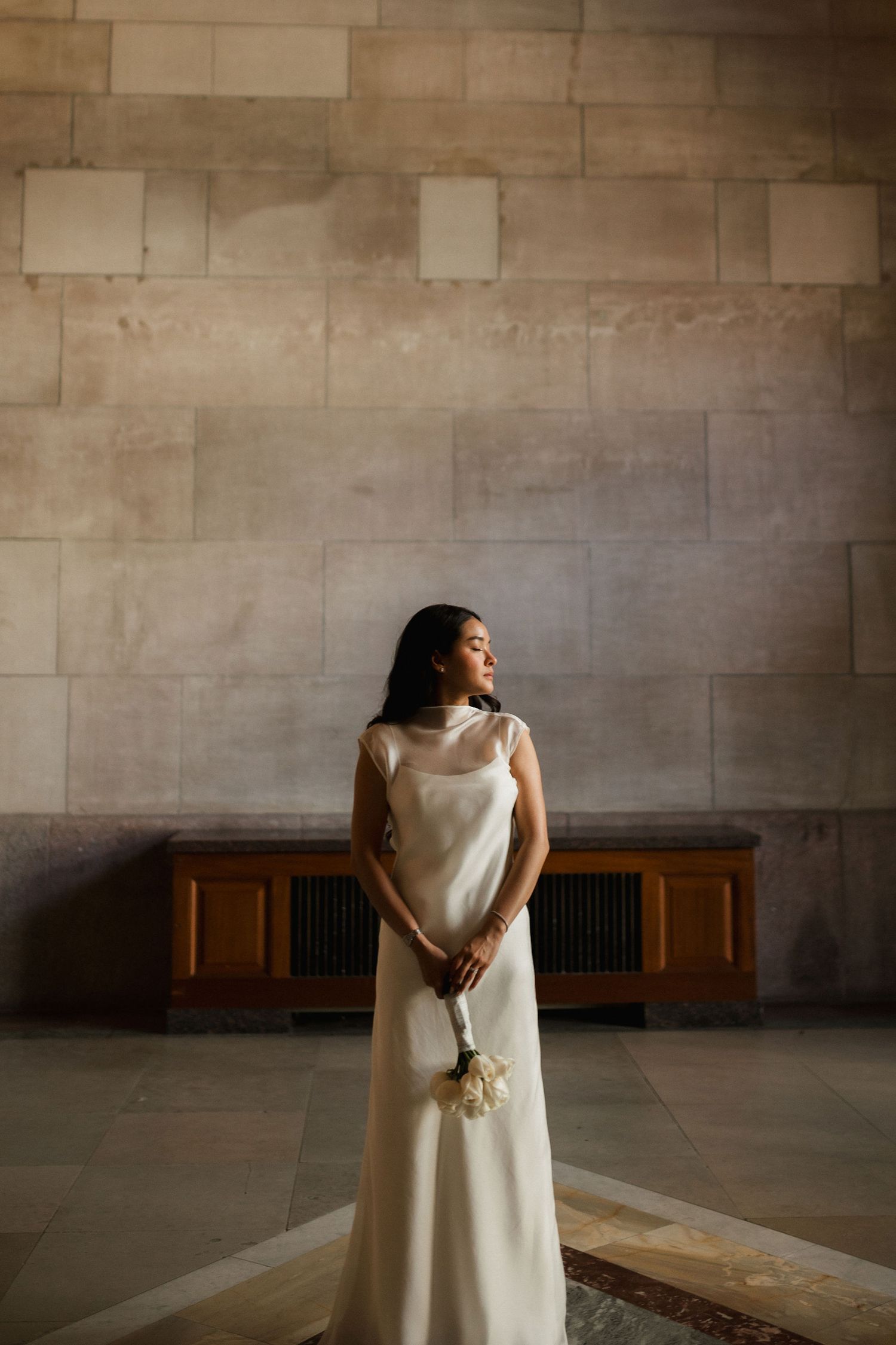 Bride in elegant white satin gown holding bouquet, standing in sunlit stone hall.