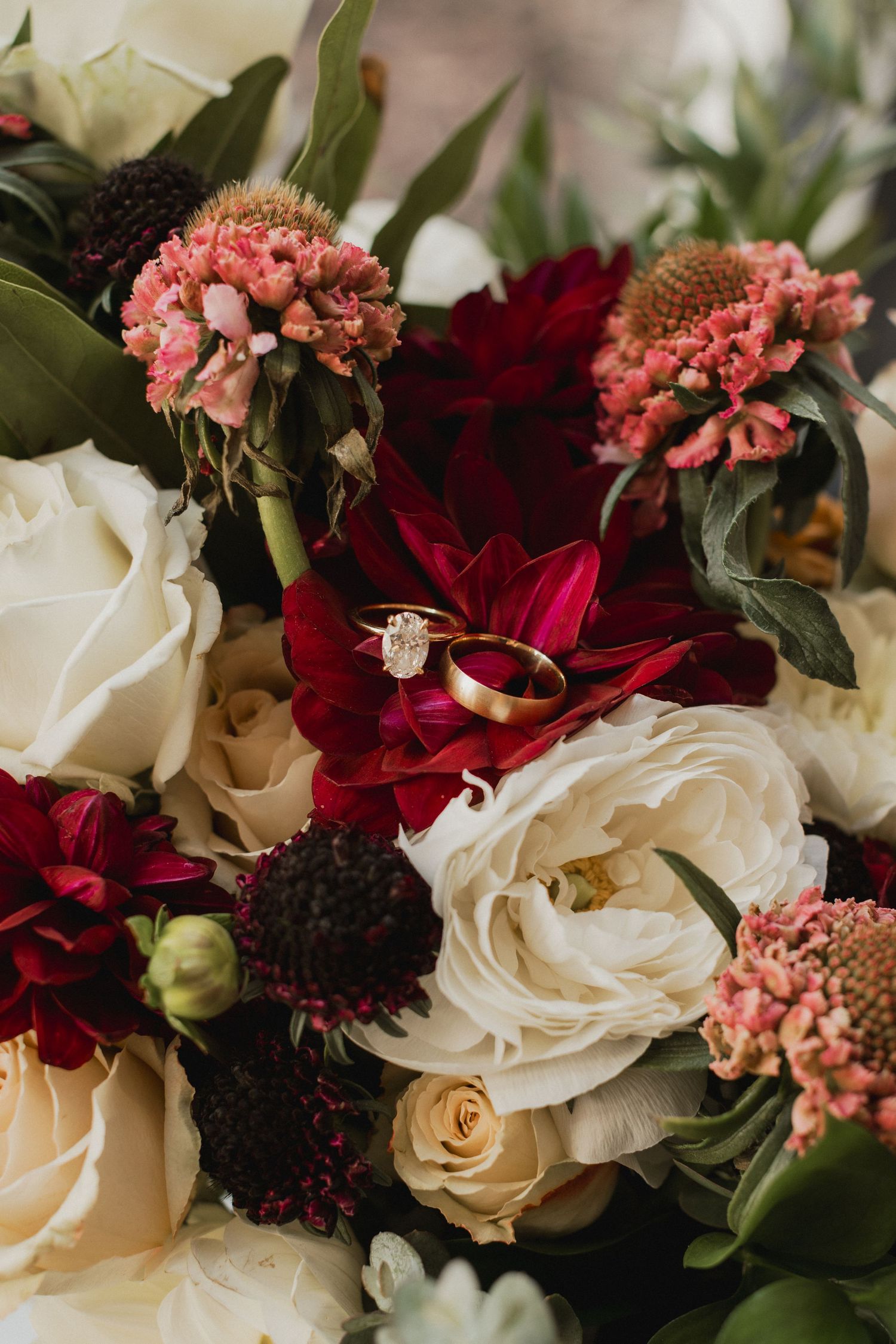 Wedding bouquet of cream roses, burgundy dahlias and pink blooms with a diamond engagement ring and gold bands resting on the petals.