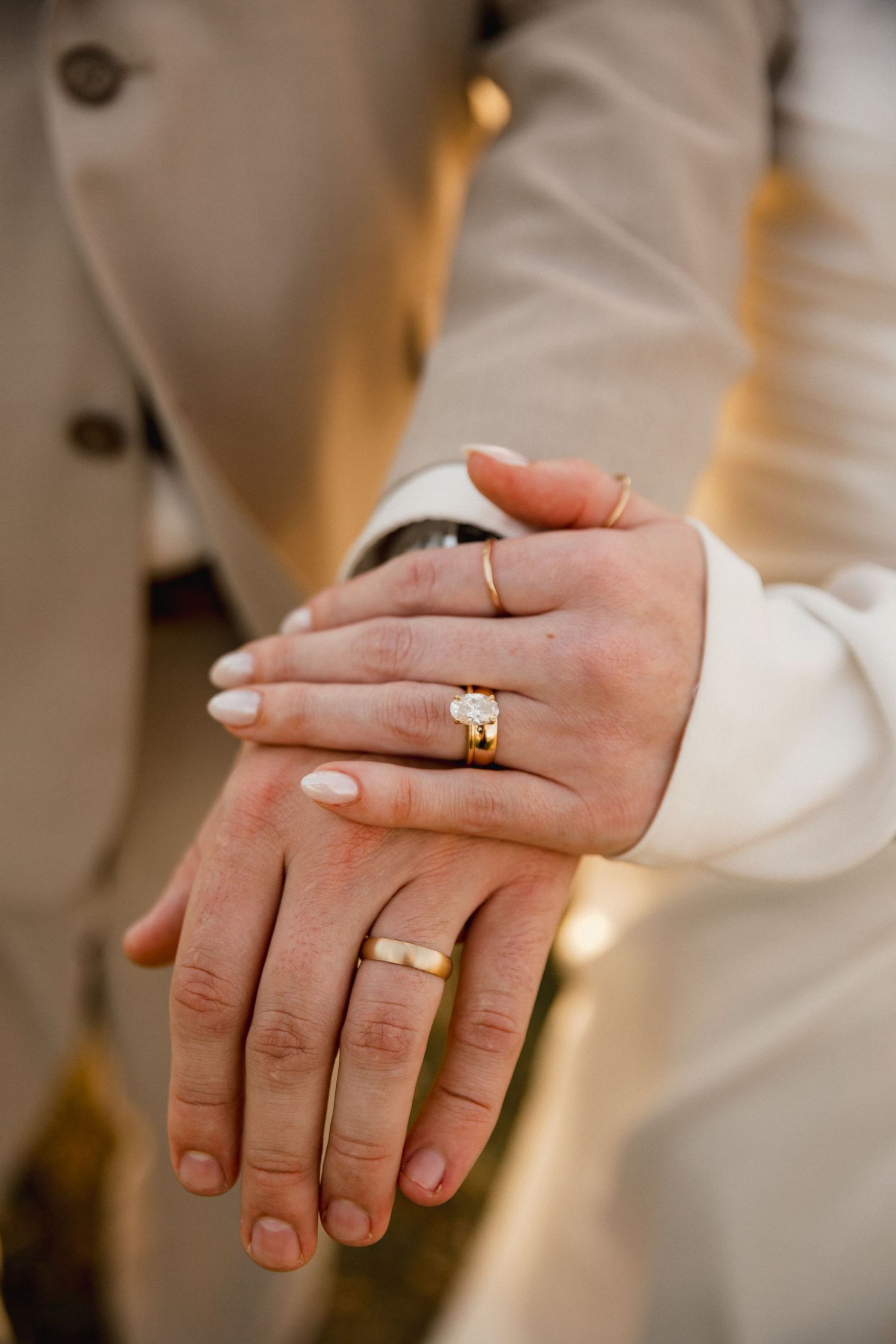 Close-up of a newlywed couple's joined hands showing gold wedding bands and a diamond engagement ring against tan wedding attire.