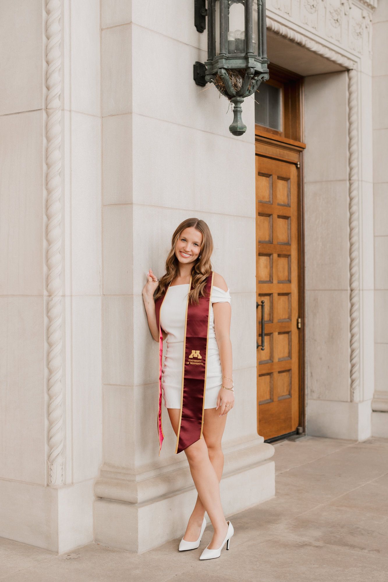 Female graduate in white dress and University of Minnesota maroon sash leaning against ornate building doorway.