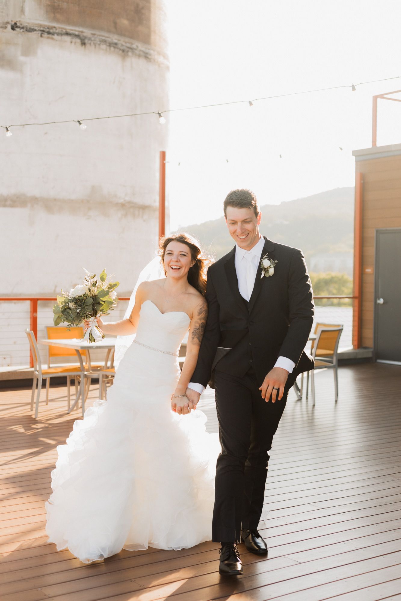 Bride and groom holding hands smiling on rooftop deck at golden hour, bride in strapless ruffle gown holding bouquet.