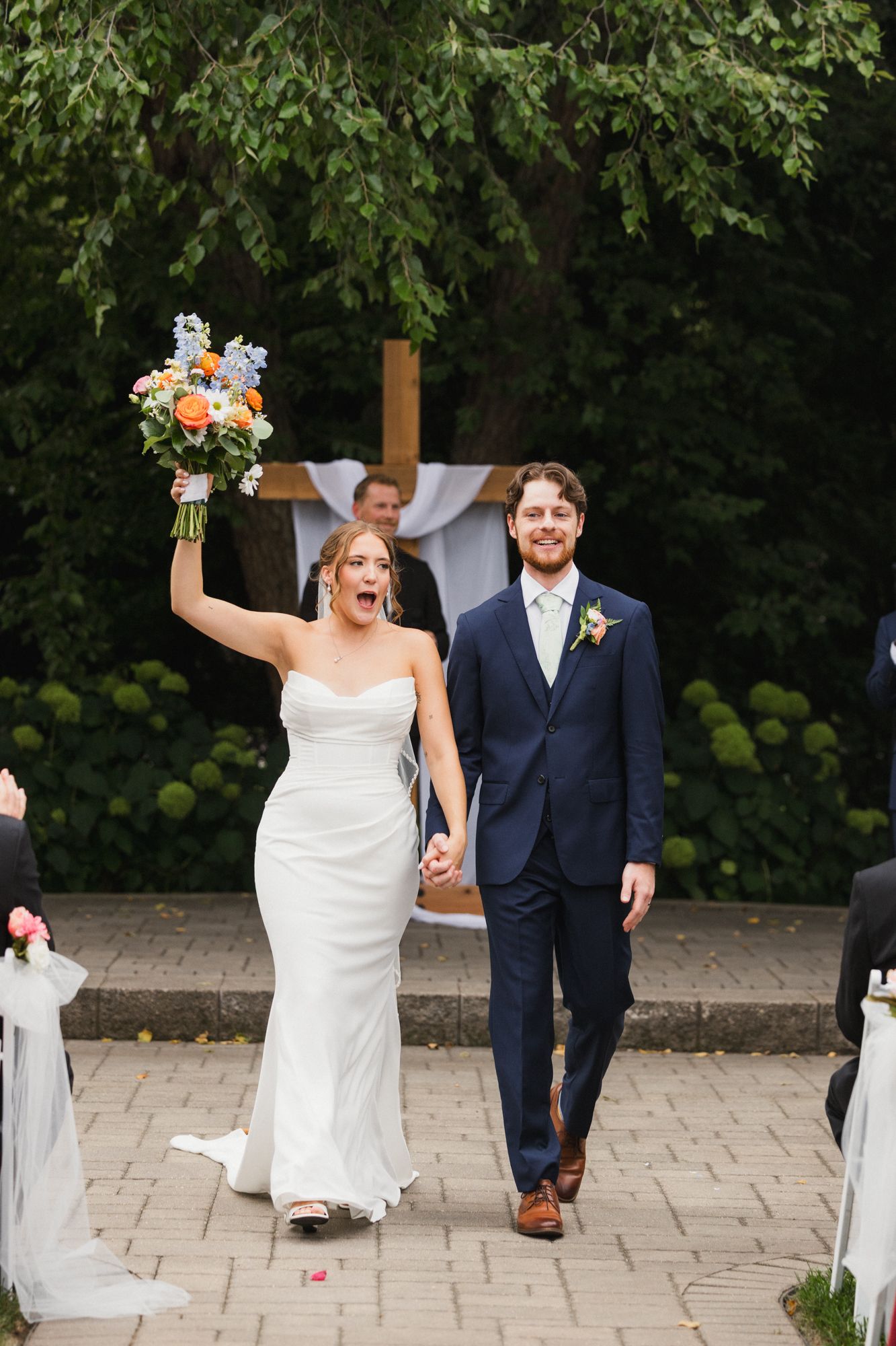 Joyful bride raises bouquet while walking down aisle with groom in navy suit after outdoor wedding ceremony.