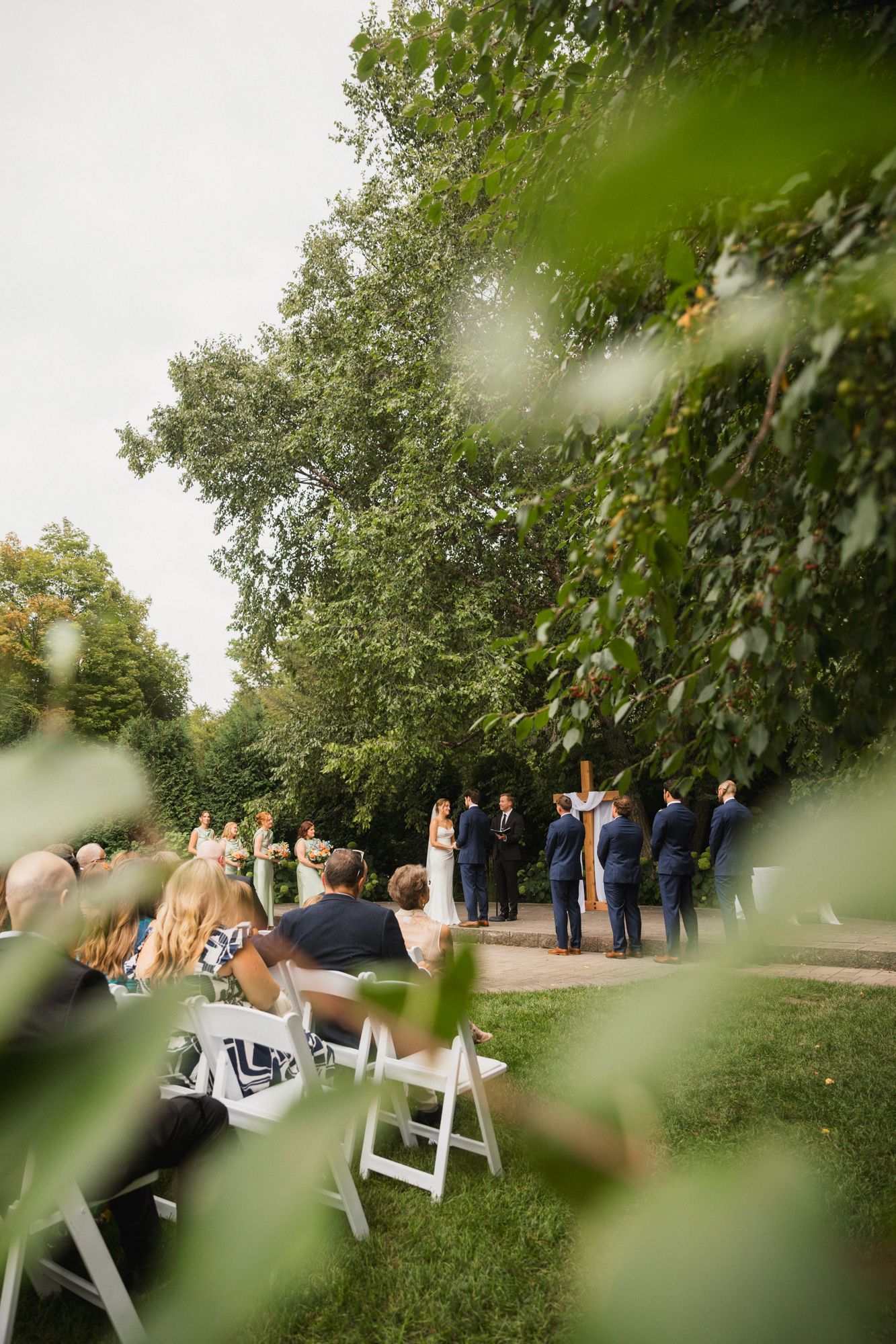 Outdoor wedding ceremony on a garden lawn with guests seated in white chairs, framed by lush green trees.