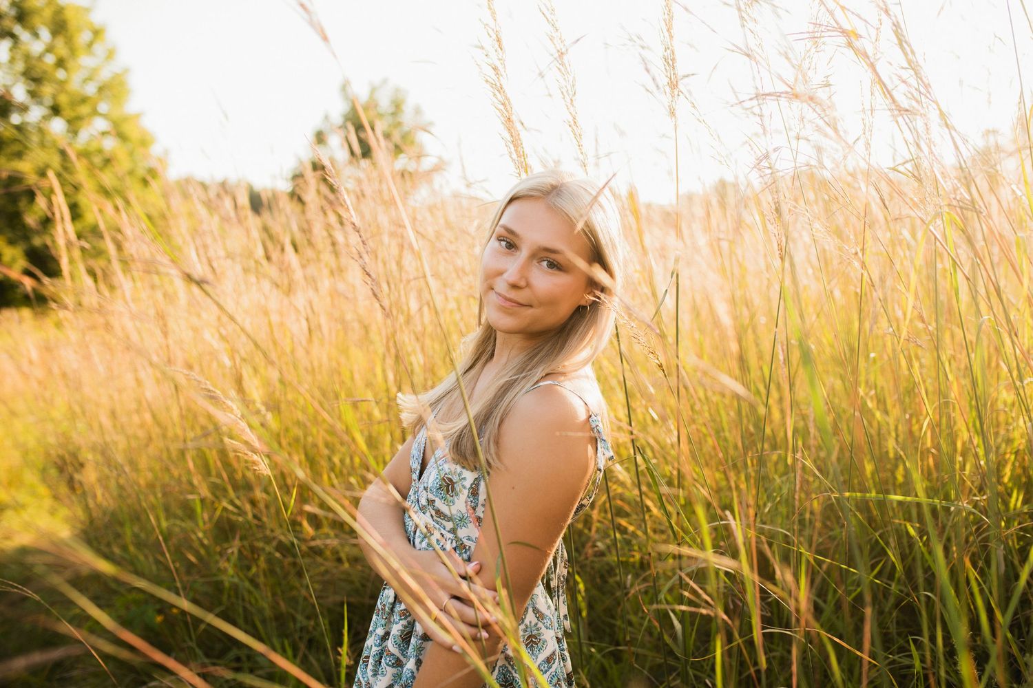 Young blonde woman in floral dress standing in golden tall grass field during sunset golden hour.