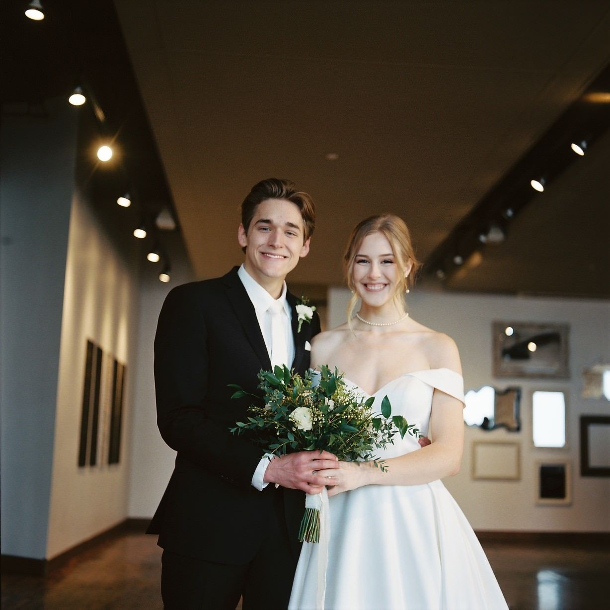 Smiling bride and groom pose together in elegant wedding attire, bride holding white and green bouquet in a gallery venue.