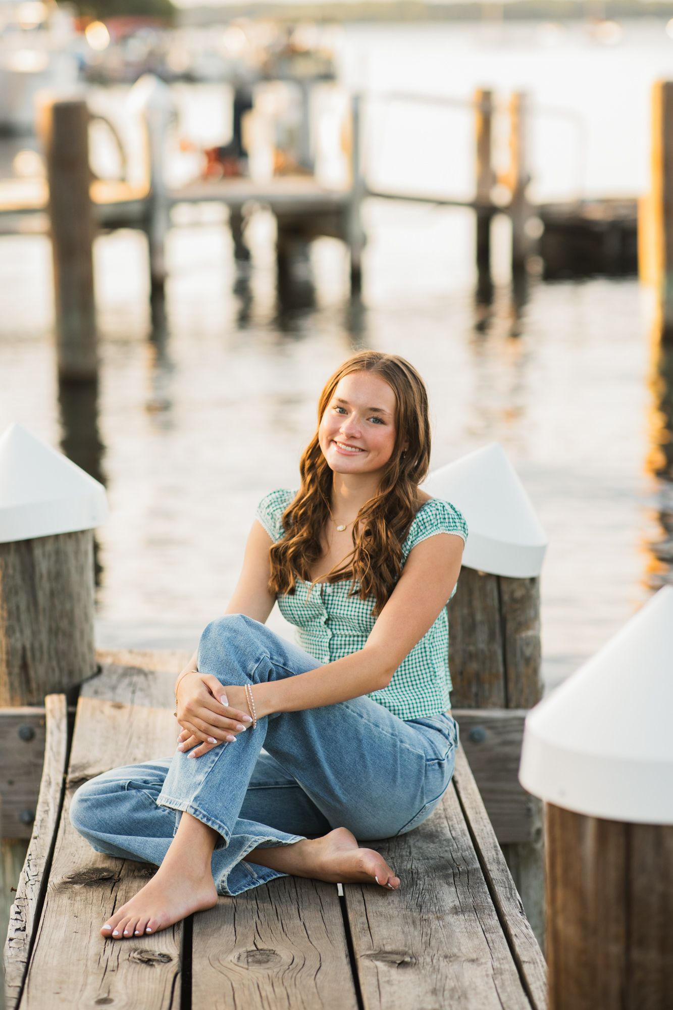 Young woman sitting cross-legged on a wooden dock by the water, wearing jeans and a green top, smiling warmly.