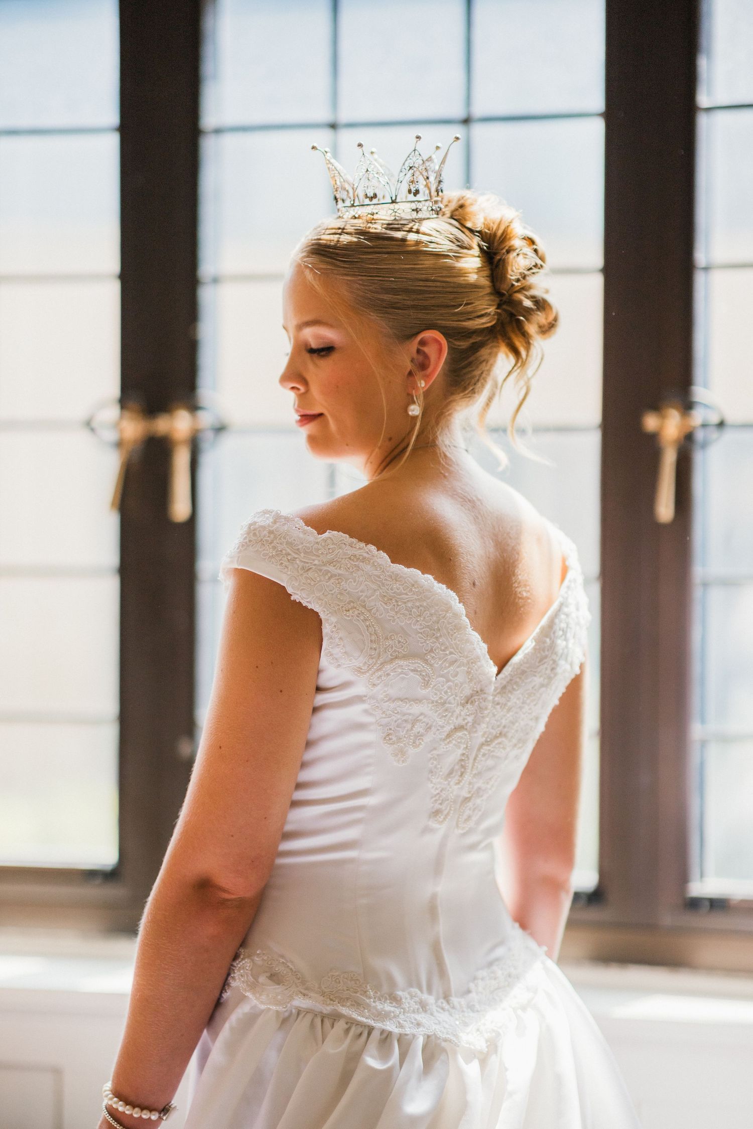 Elegant bride in white lace off-shoulder gown wearing a sparkling tiara, photographed from behind near tall windows.