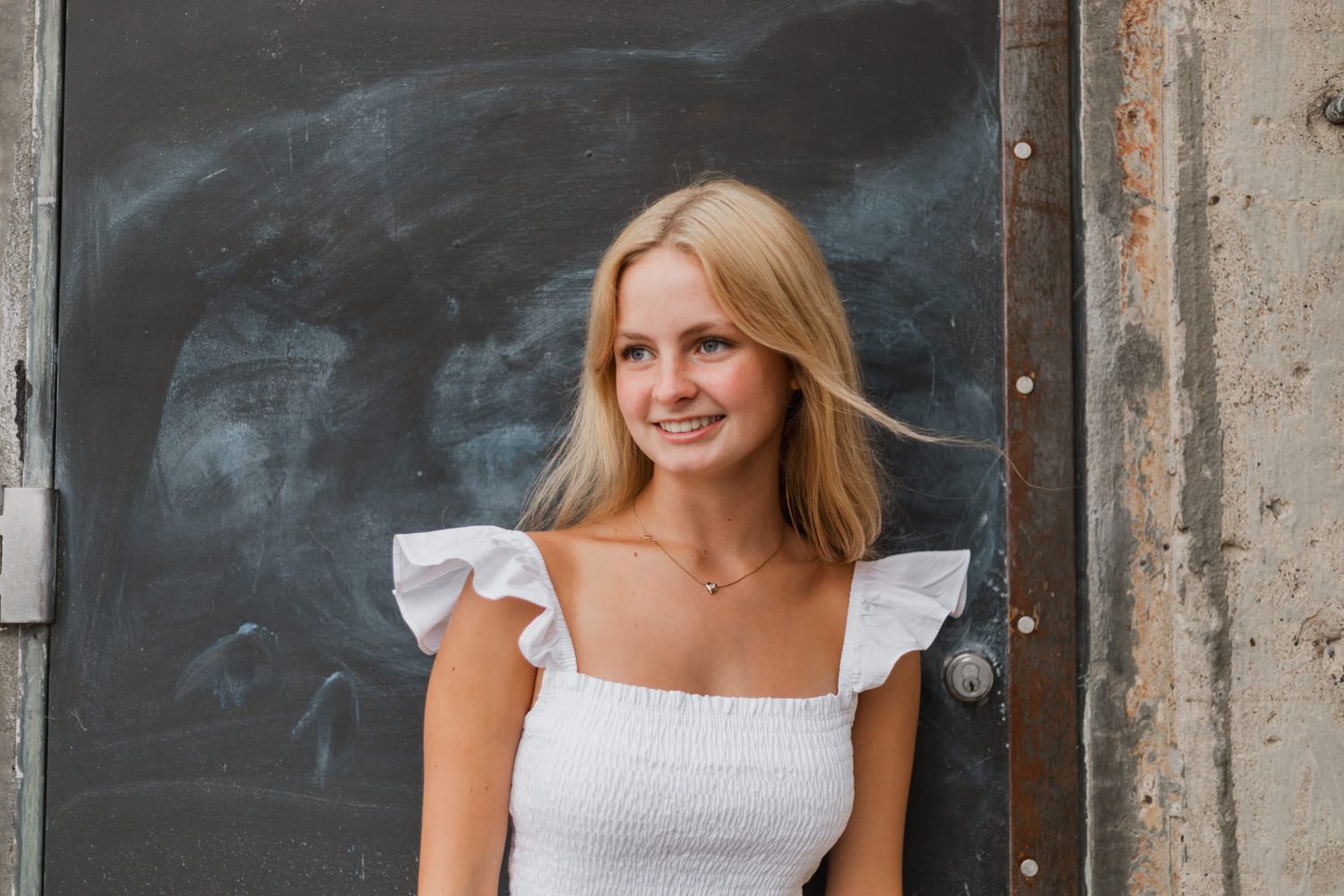 A woman in a white dress smiles while standing against a rustic stone wall background.