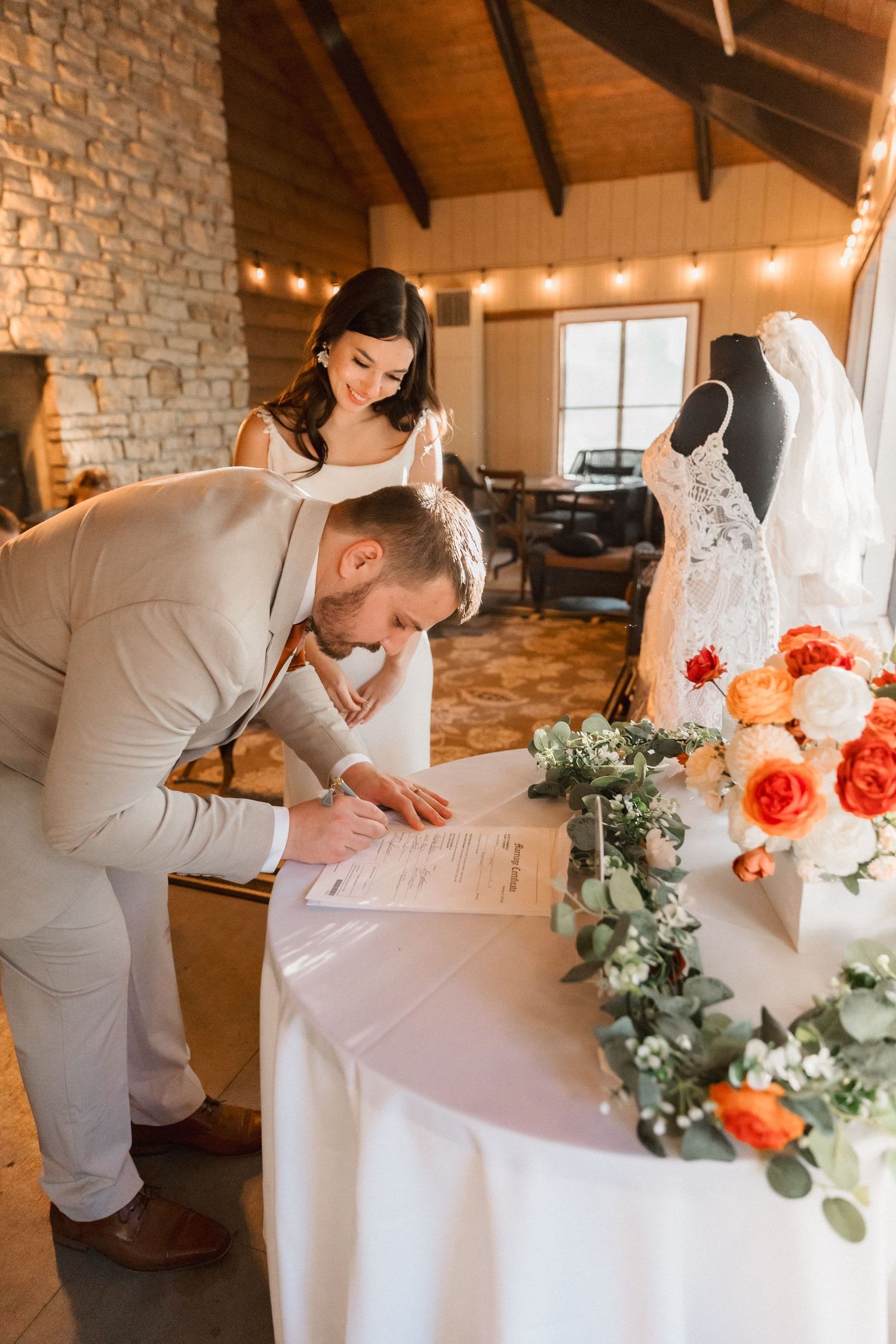 A bride signs wedding documents at a white table with coral flowers and greenery in a rustic stone venue.