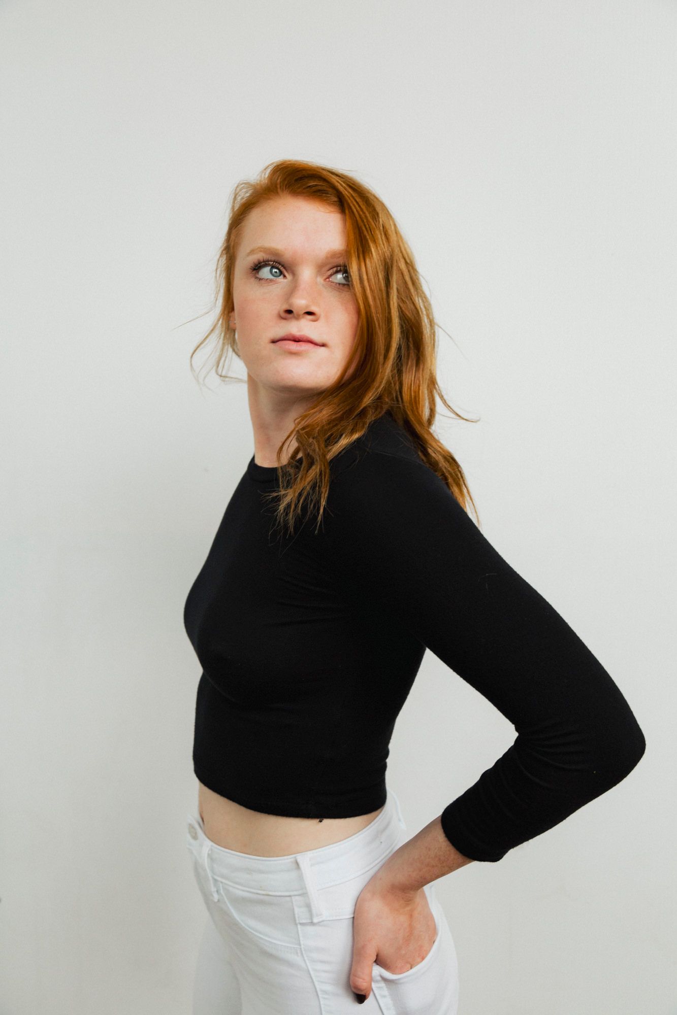 Stylish studio portrait of a redhead in a black crop top and white pants against a white background.