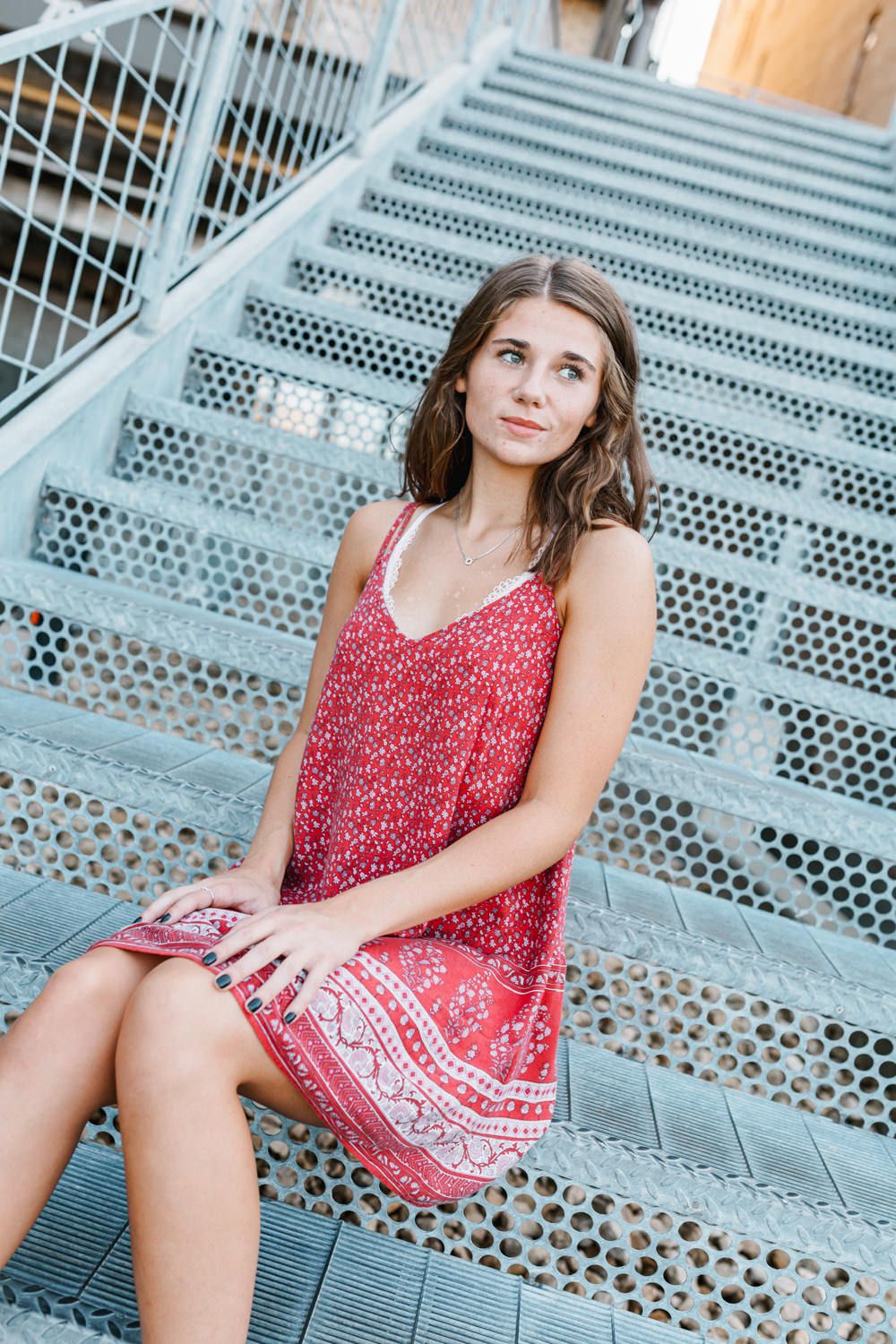 Young woman in red patterned sundress sitting on industrial metal stairs.