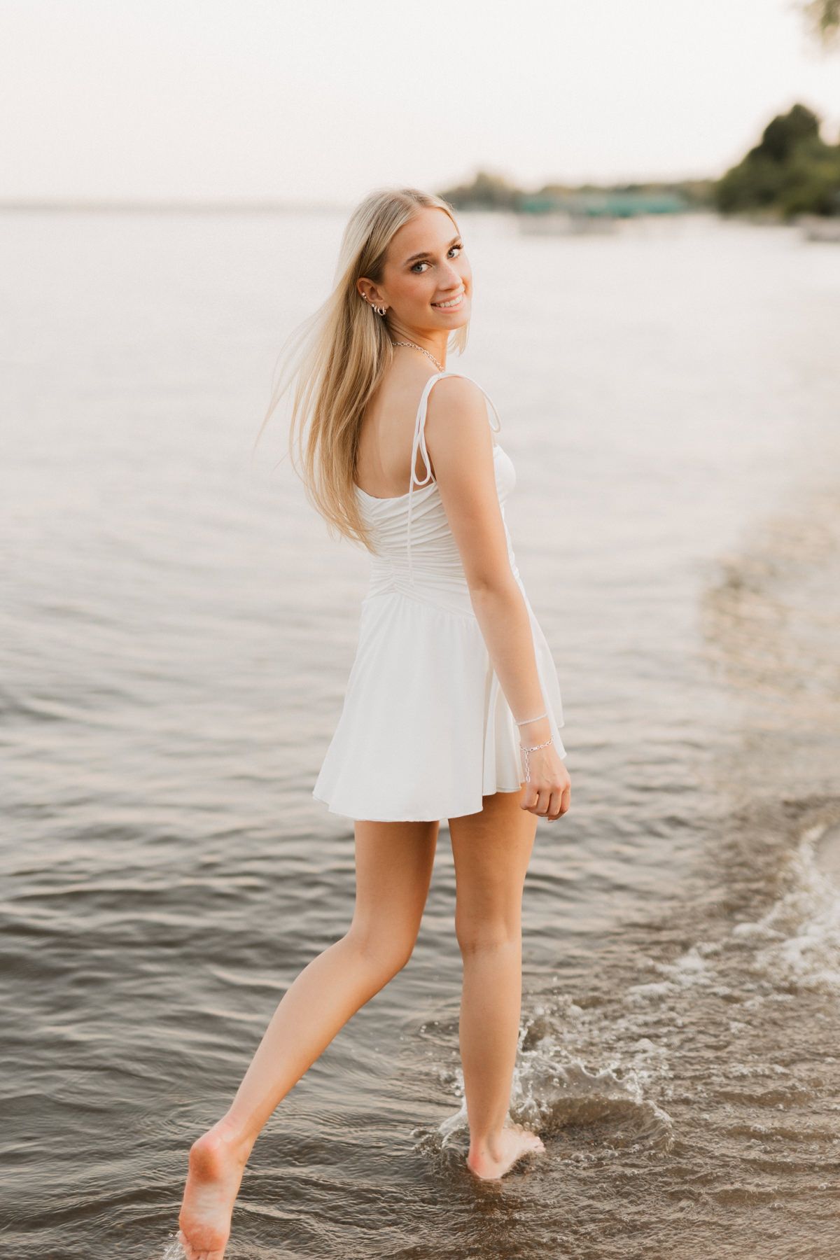 Young blonde woman in white mini dress wading in ocean waves at sunset, smiling over her shoulder.