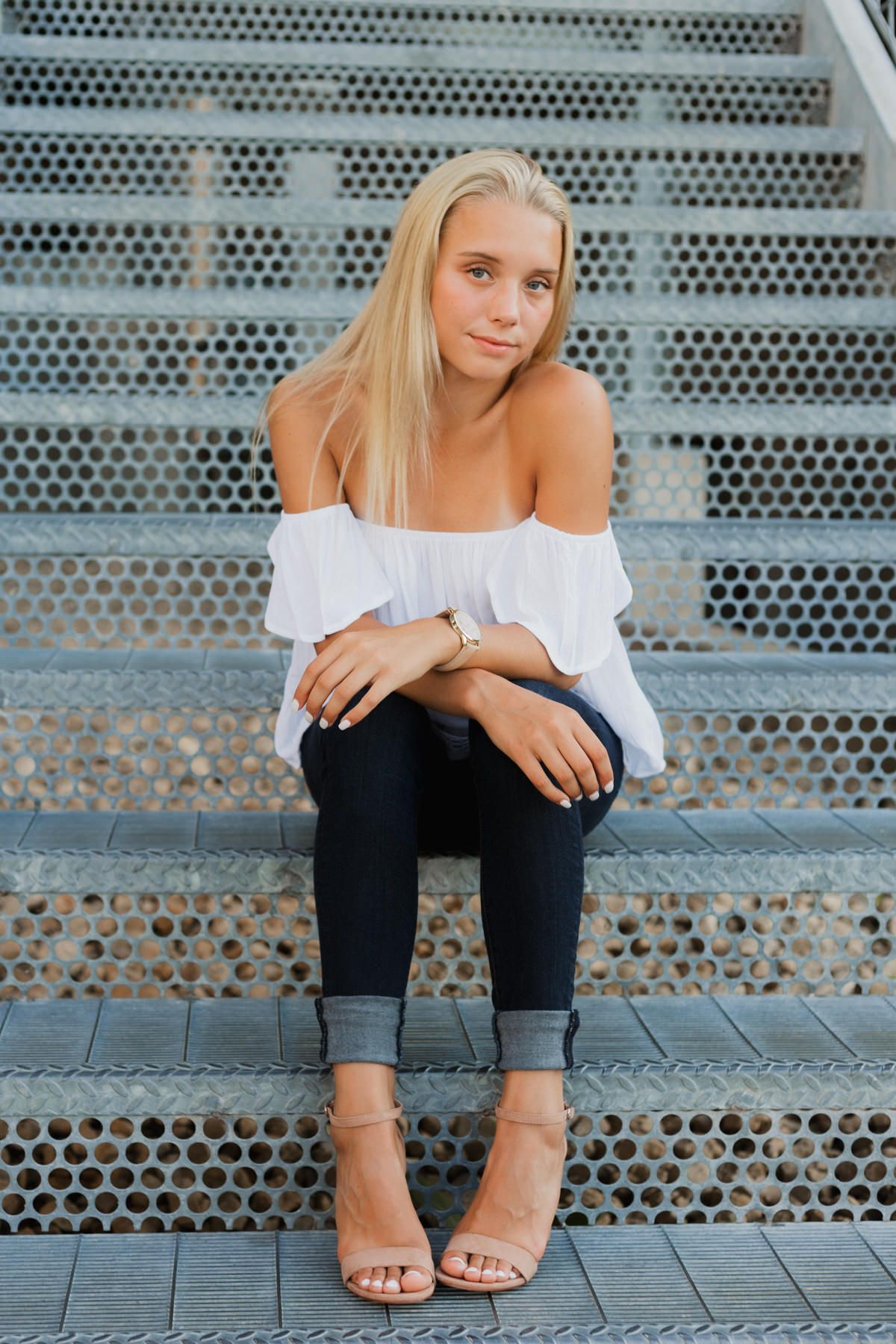 Blonde teen girl in white off-shoulder top and jeans sitting on metal perforated steps wearing heeled sandals.