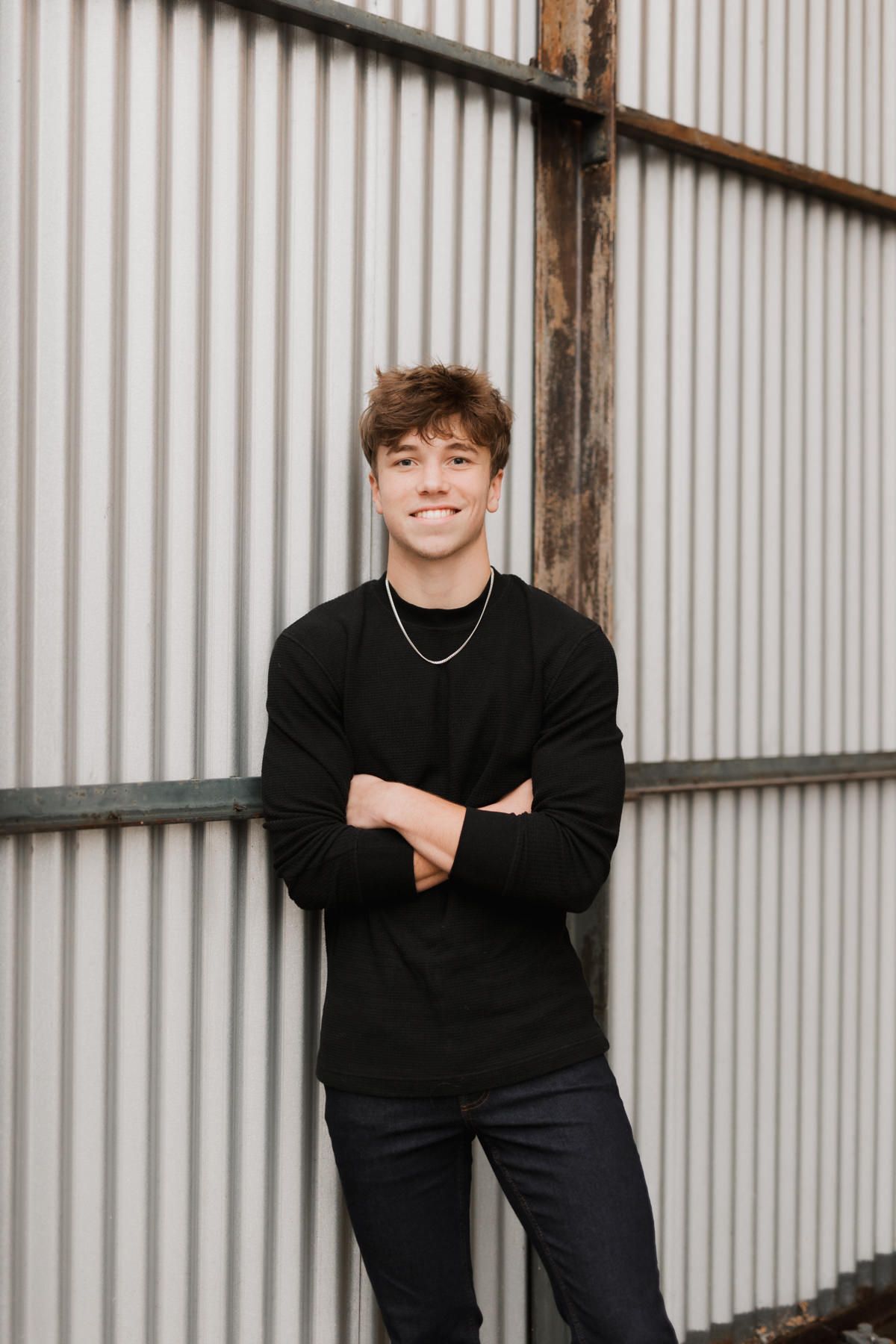 Smiling young man in black long sleeve shirt with arms crossed leaning against corrugated metal wall.