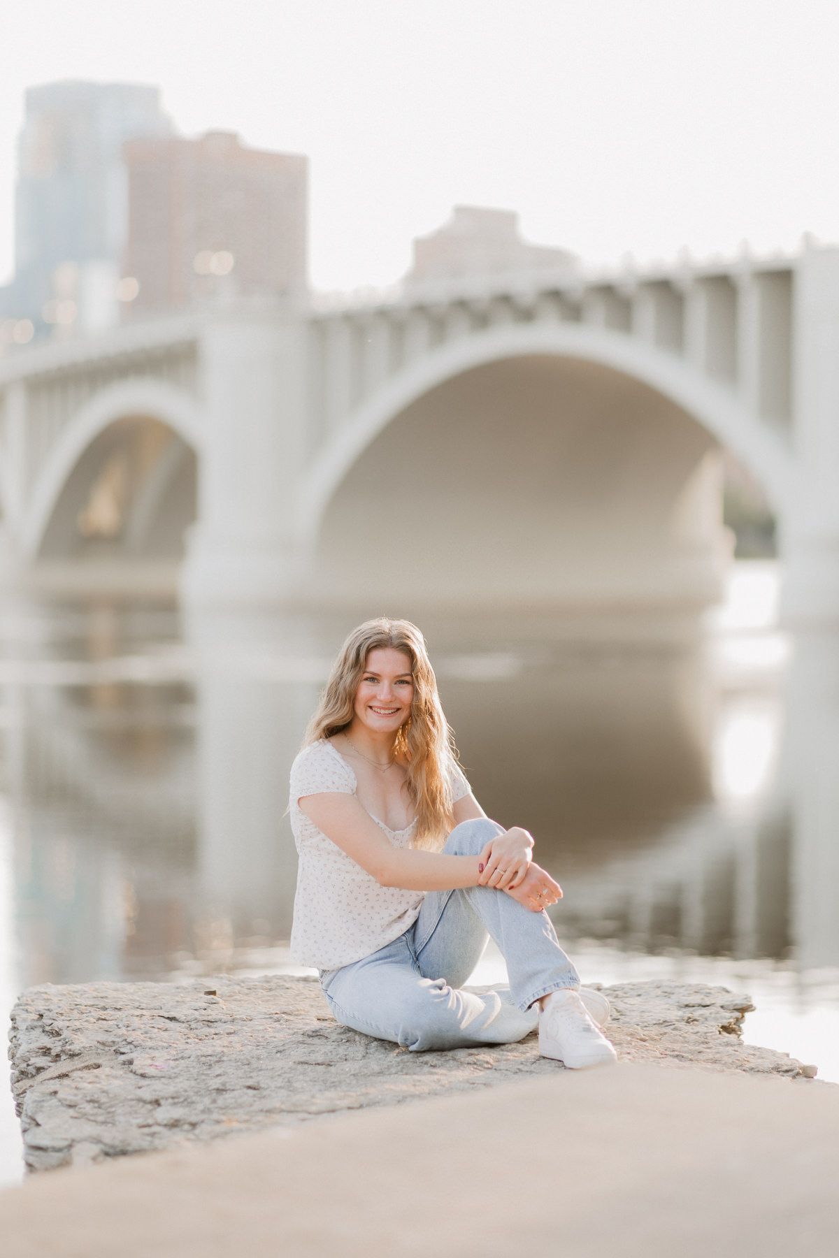 Smiling blonde woman in jeans sitting on rocks by a river with an arched bridge in the background.