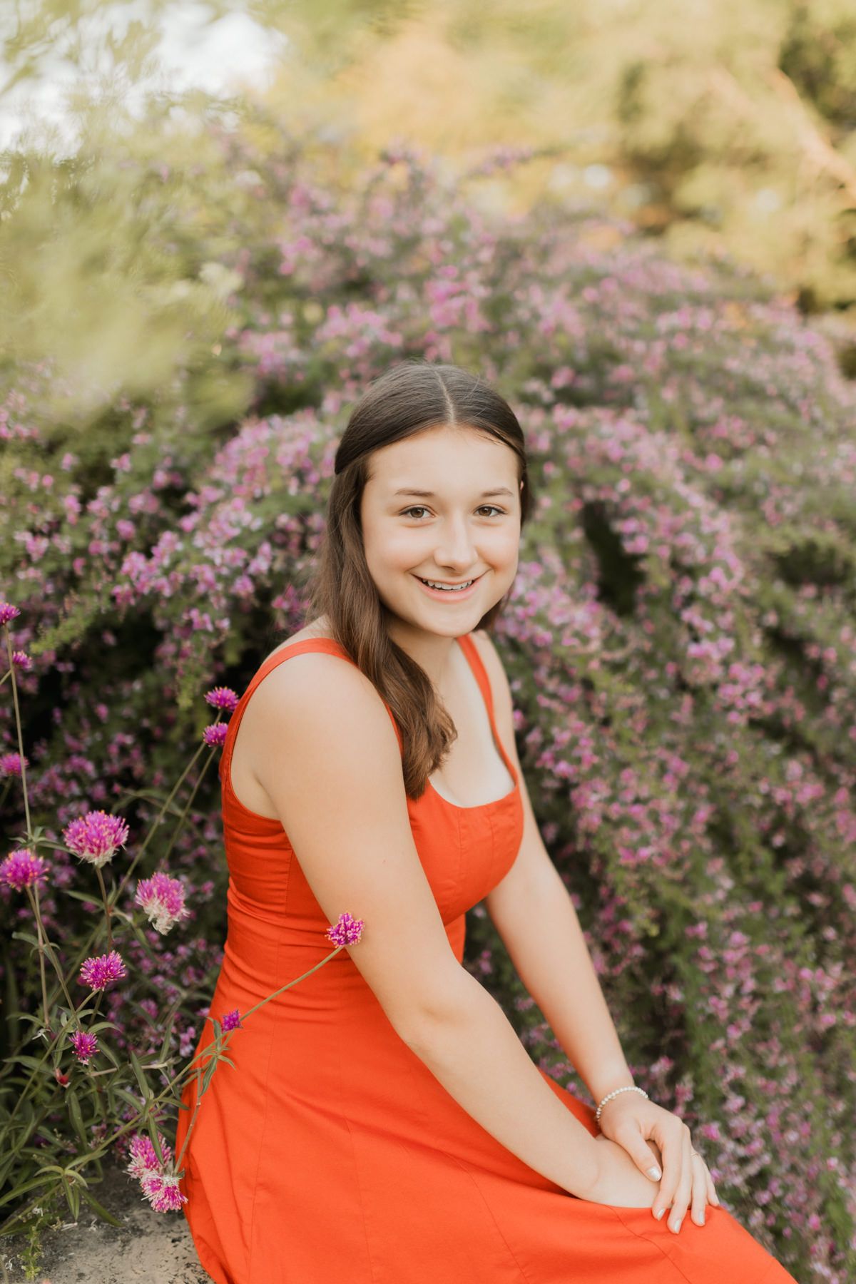 Smiling teen girl in orange dress sitting among vibrant purple flowering bushes in garden setting.
