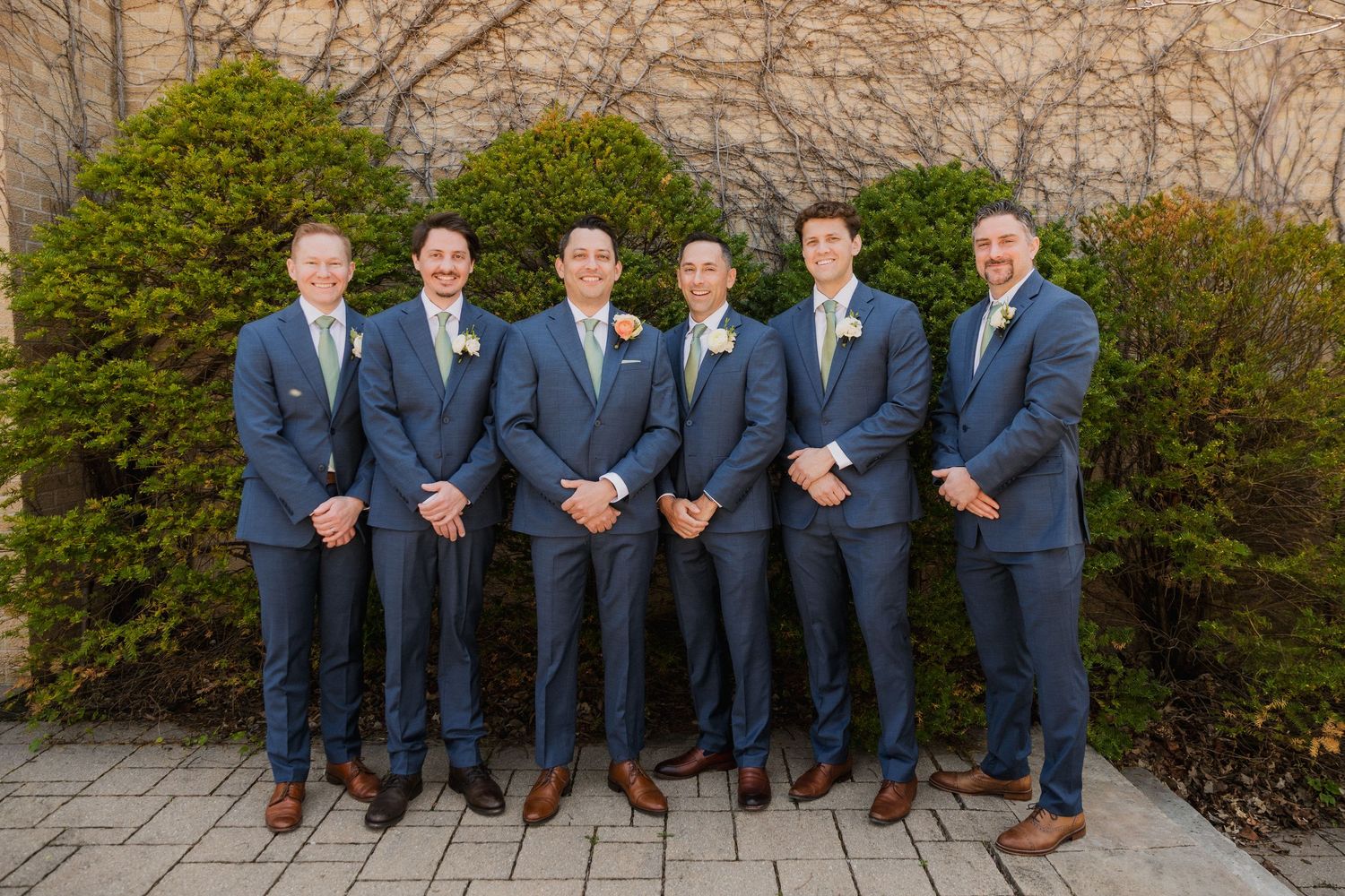 Six groomsmen in matching navy blue suits with boutonnieres pose outdoors on a brick patio for a wedding photo.