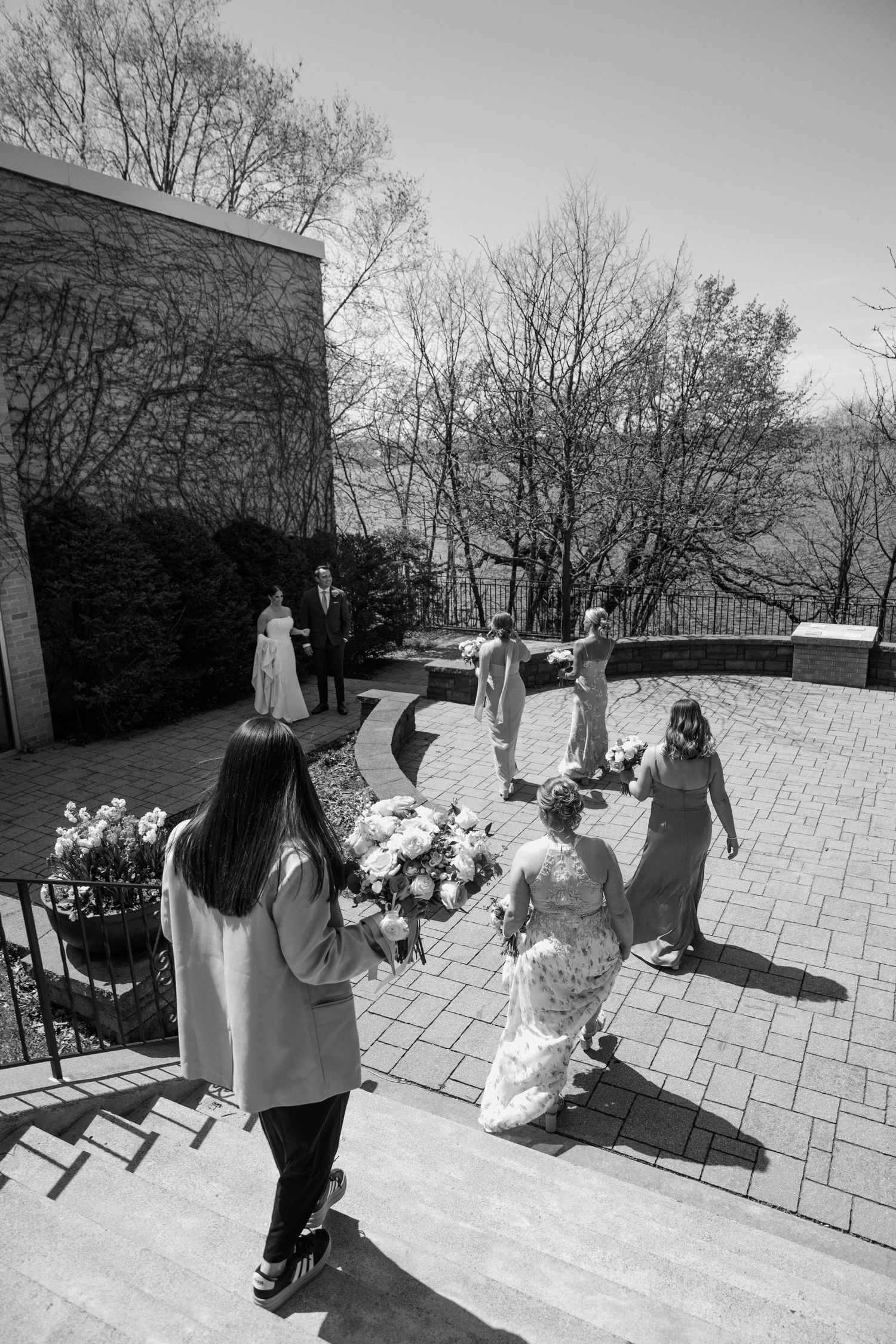 Black and white photo of a bride and bridesmaids walking on a brick patio toward the groom on a sunny day.