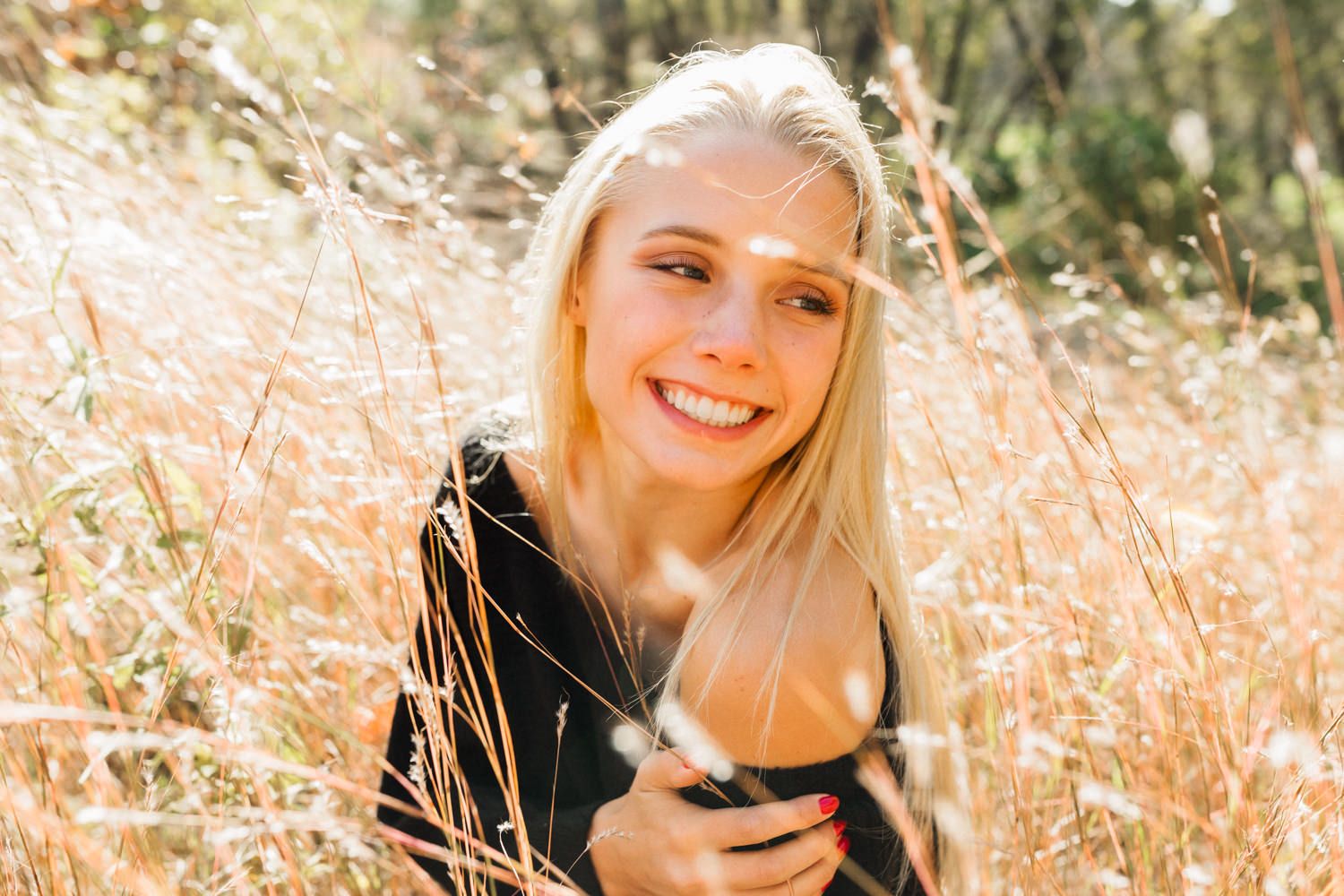 Smiling blonde woman sitting in golden field grass, holding a camera, bathed in warm sunlight outdoors.
