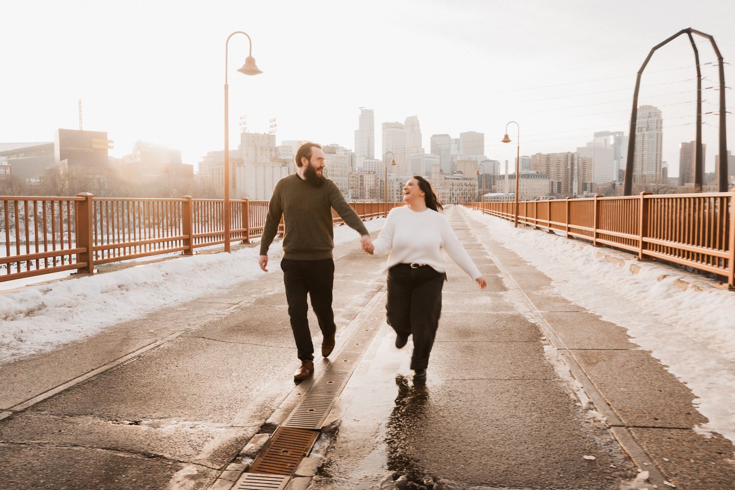 Two people walk hand in hand along a bridge pathway at sunset with an urban skyline in the distance.