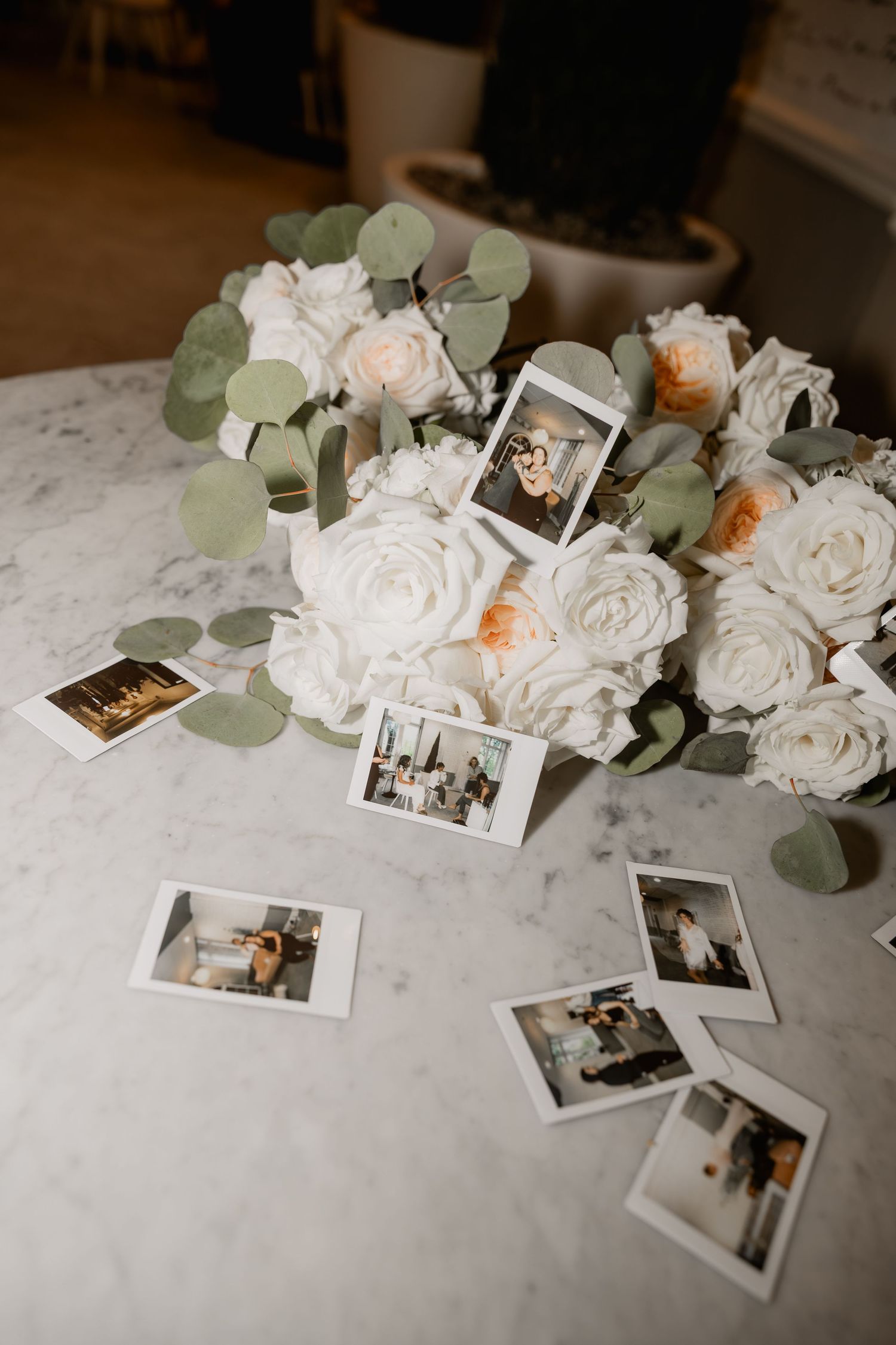 White roses and eucalyptus bouquet on marble table with scattered Polaroid photos at a wedding reception.