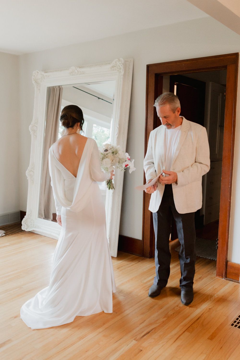 A person in a white wedding gown stands before a large ornate mirror while another in a white jacket looks on.