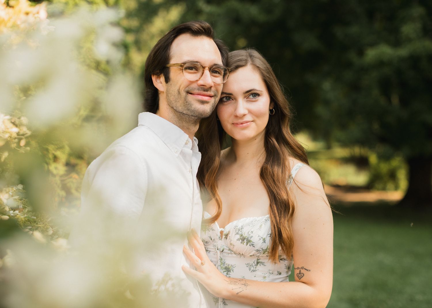 Couple smiling together outdoors surrounded by blooming white flowers in a lush green garden setting.