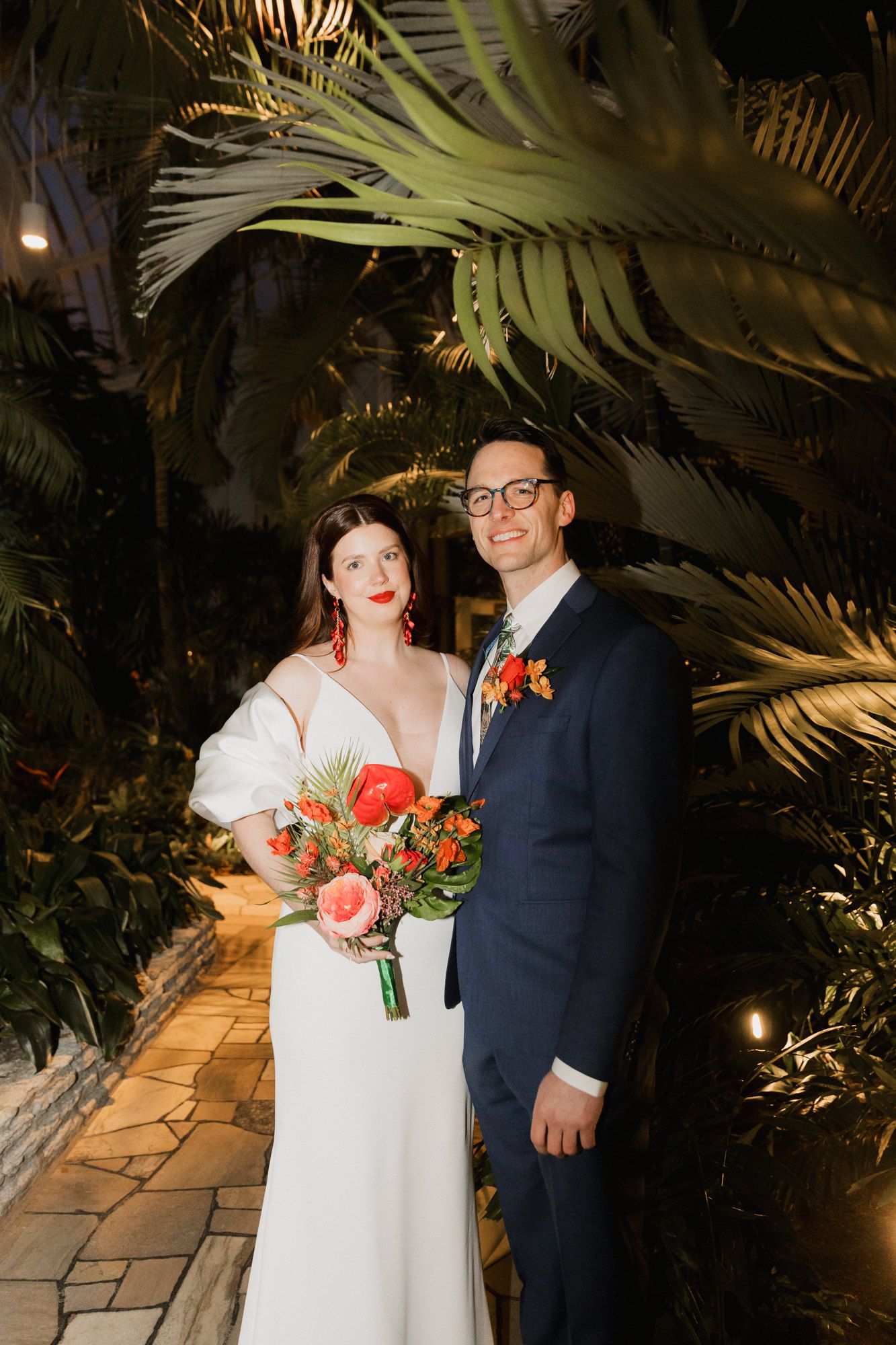 Bride in white gown with bouquet and groom in navy suit pose among tropical palms at night wedding.