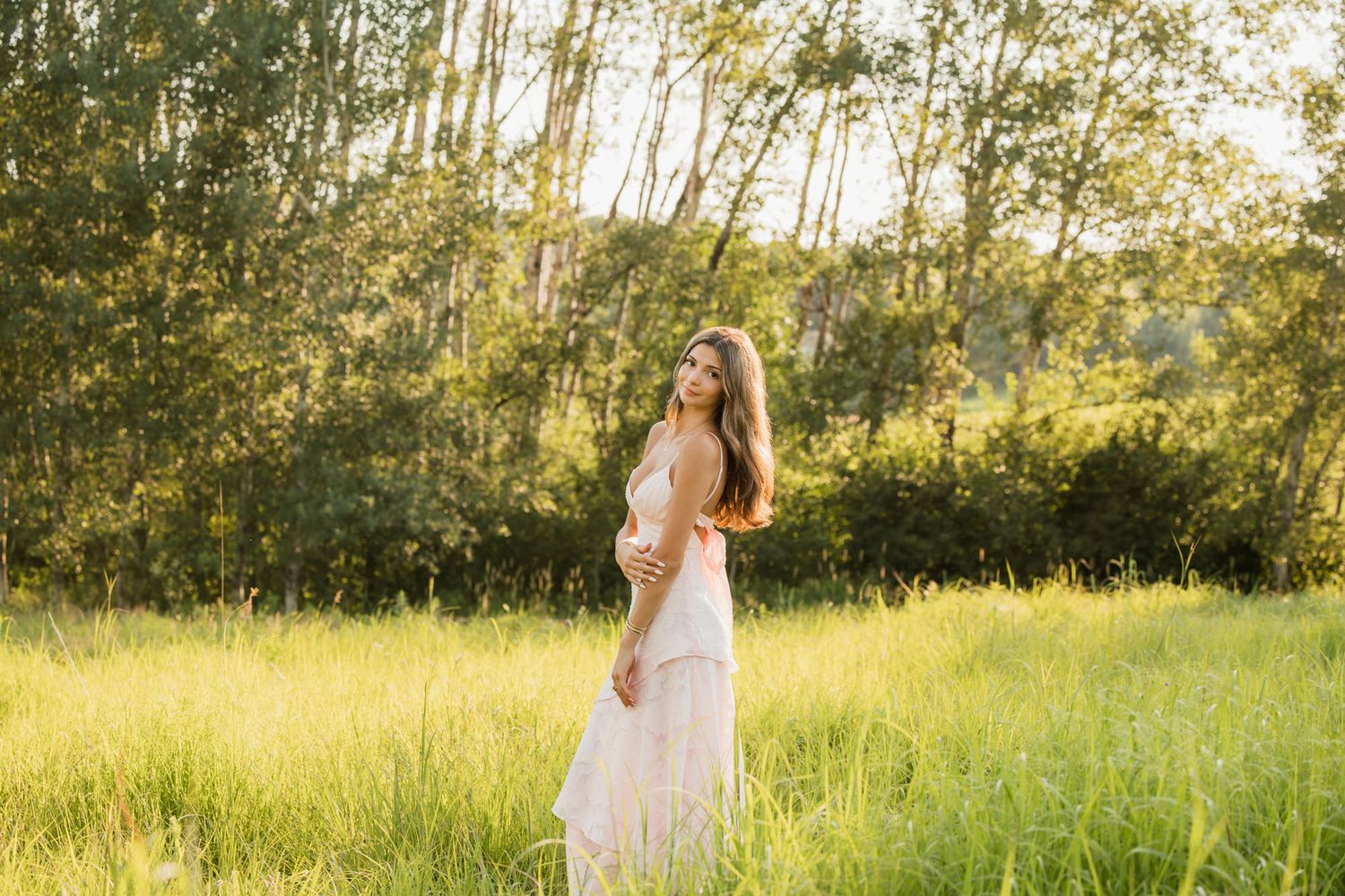 Elegant woman in a white maxi dress standing in a sunlit meadow surrounded by tall green grass and trees.